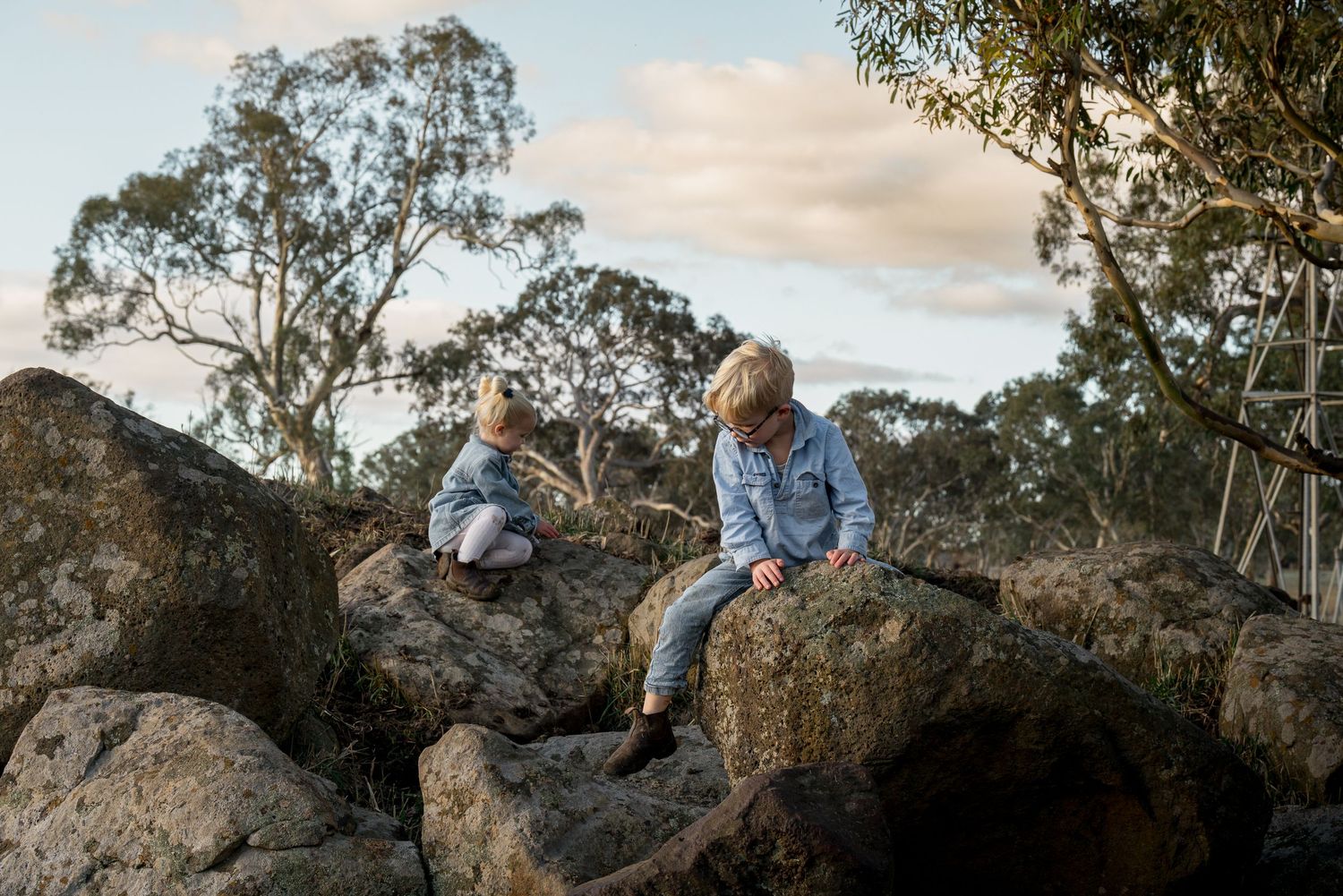 Two children in denim jackets climbing and sitting on large rocks surrounded by eucalyptus trees outdoors.