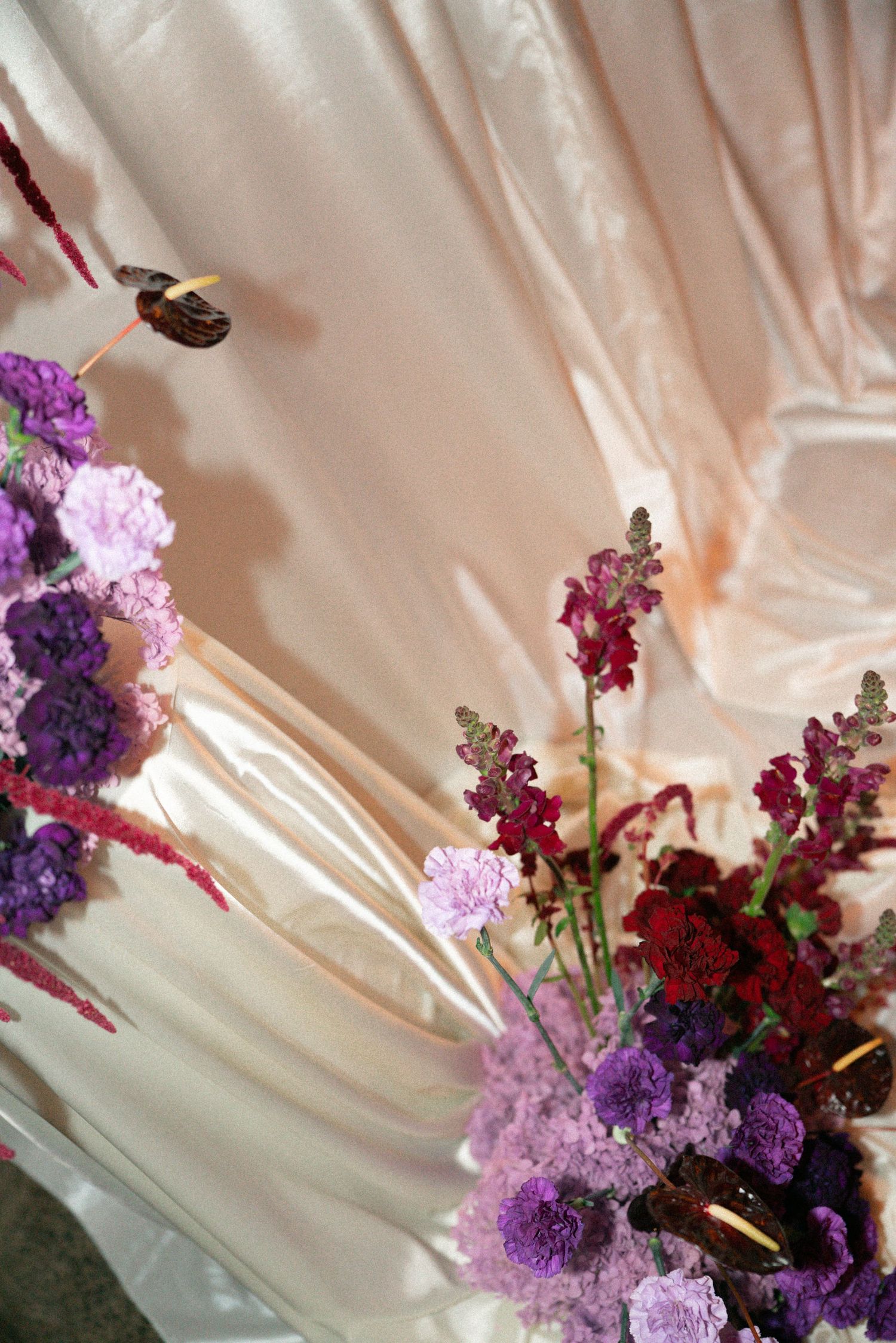 Purple and burgundy floral arrangement with carnations against draped pink satin fabric backdrop.
