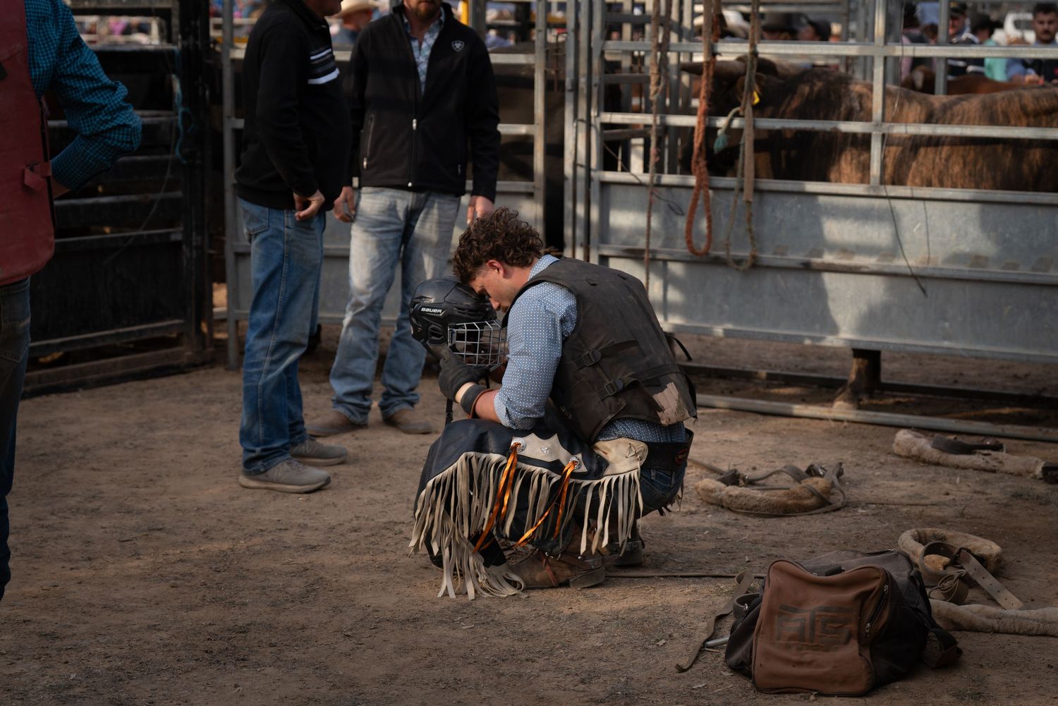 Cowboy crouching down adjusting fringed leather chaps near a rodeo arena with spectators nearby.