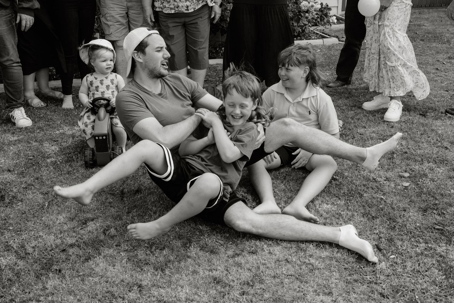 Father sitting on grass laughing with two young children at an outdoor family gathering.