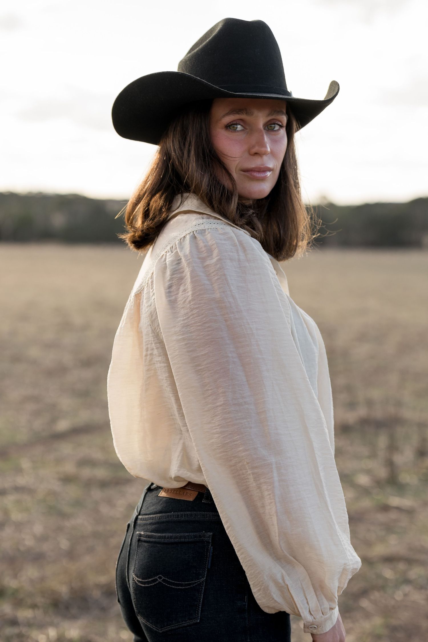 Brunette woman wearing black cowboy hat and ivory blouse posing in an open field at sunset.