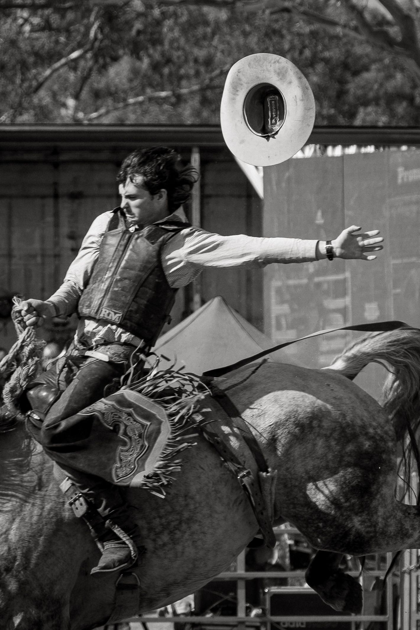 Black and white photo of a rodeo cowboy riding a bucking horse while his hat flies off.
