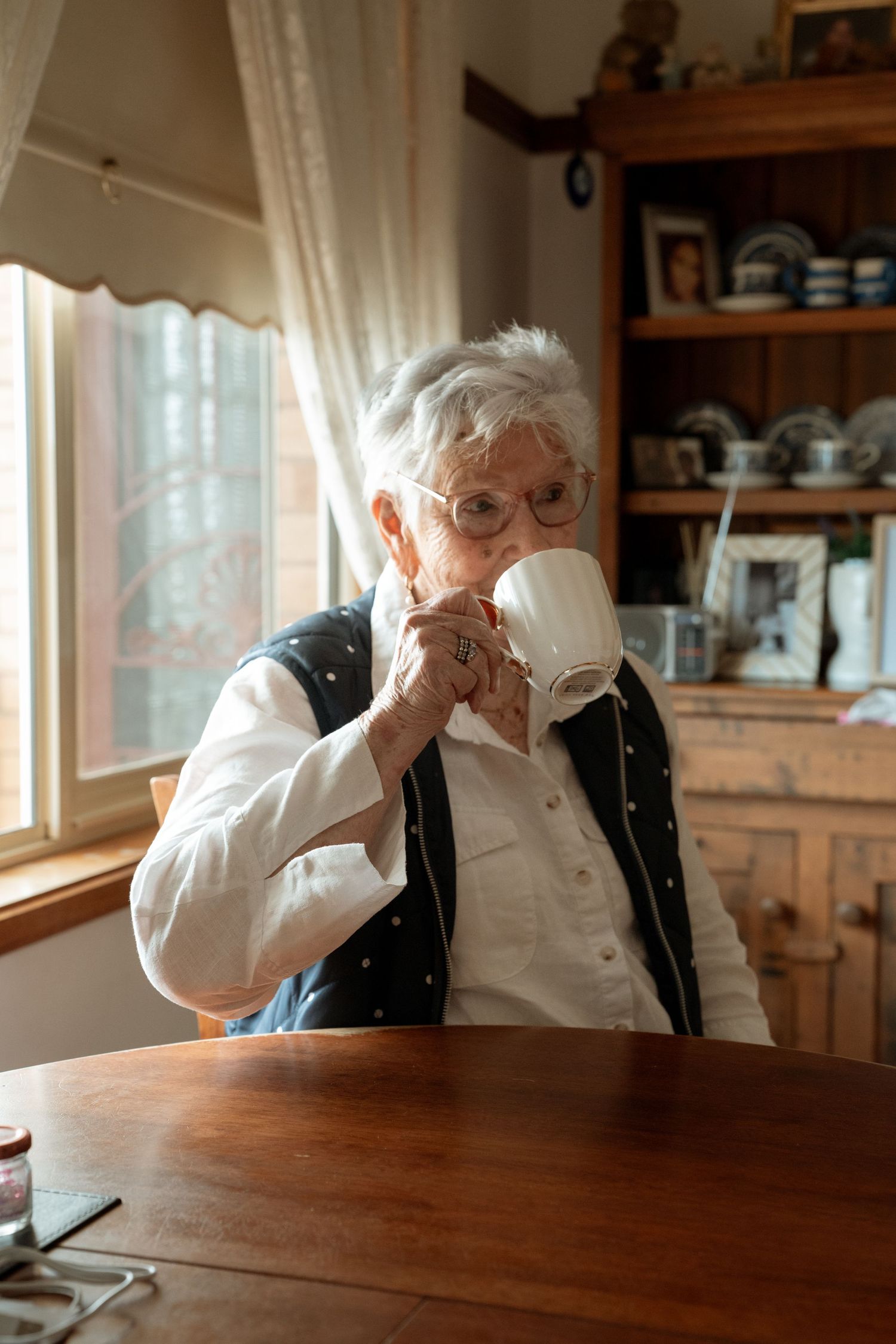 Elderly white-haired woman drinking from a white mug at a wooden table by a sunny window.