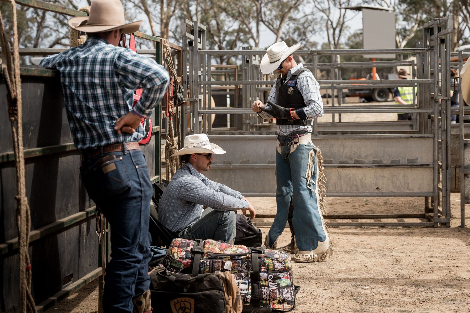 Three cowboys in jeans and hats preparing rodeo gear beside metal livestock pens.
