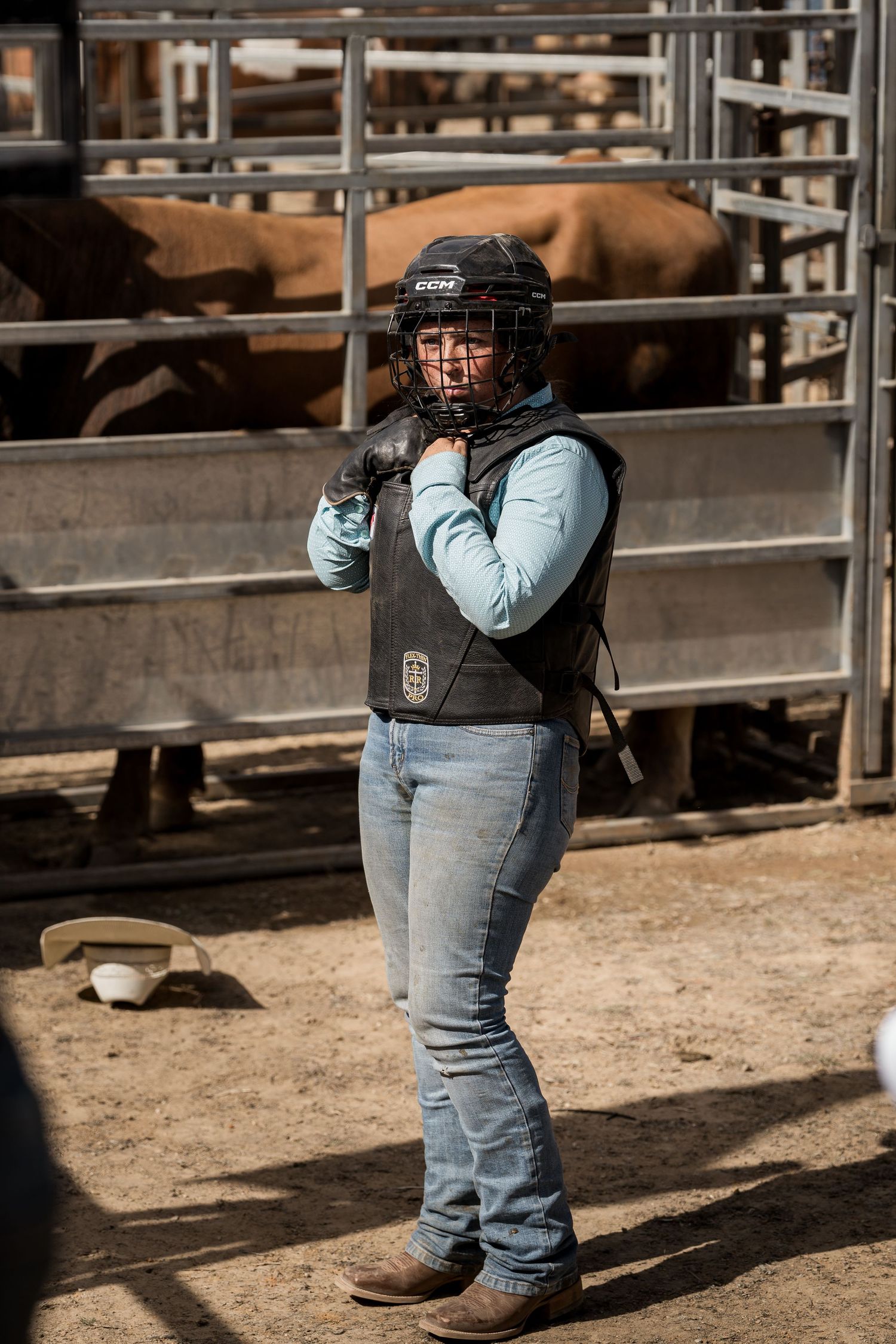 Female rodeo rider in protective helmet and vest standing near livestock pens preparing to compete.