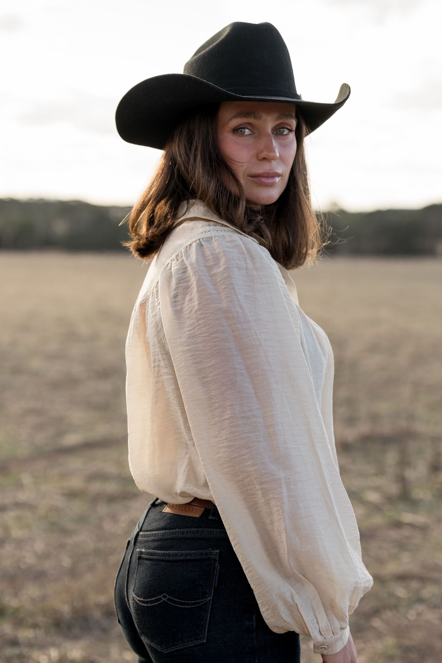 Woman in black cowboy hat and cream blouse standing in an open field at dusk.