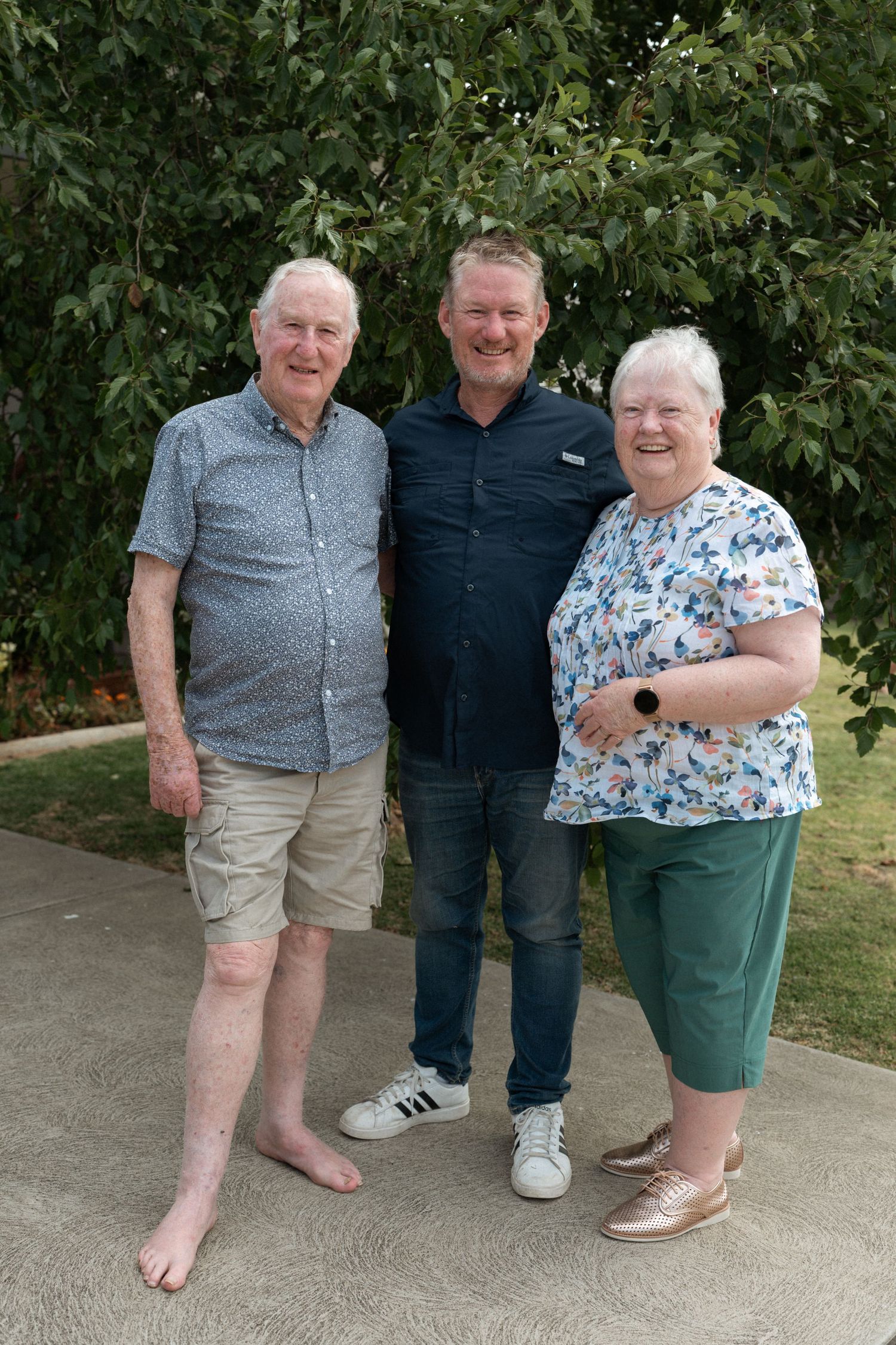Three generations of family smiling together outdoors in a garden setting on a sunny day.