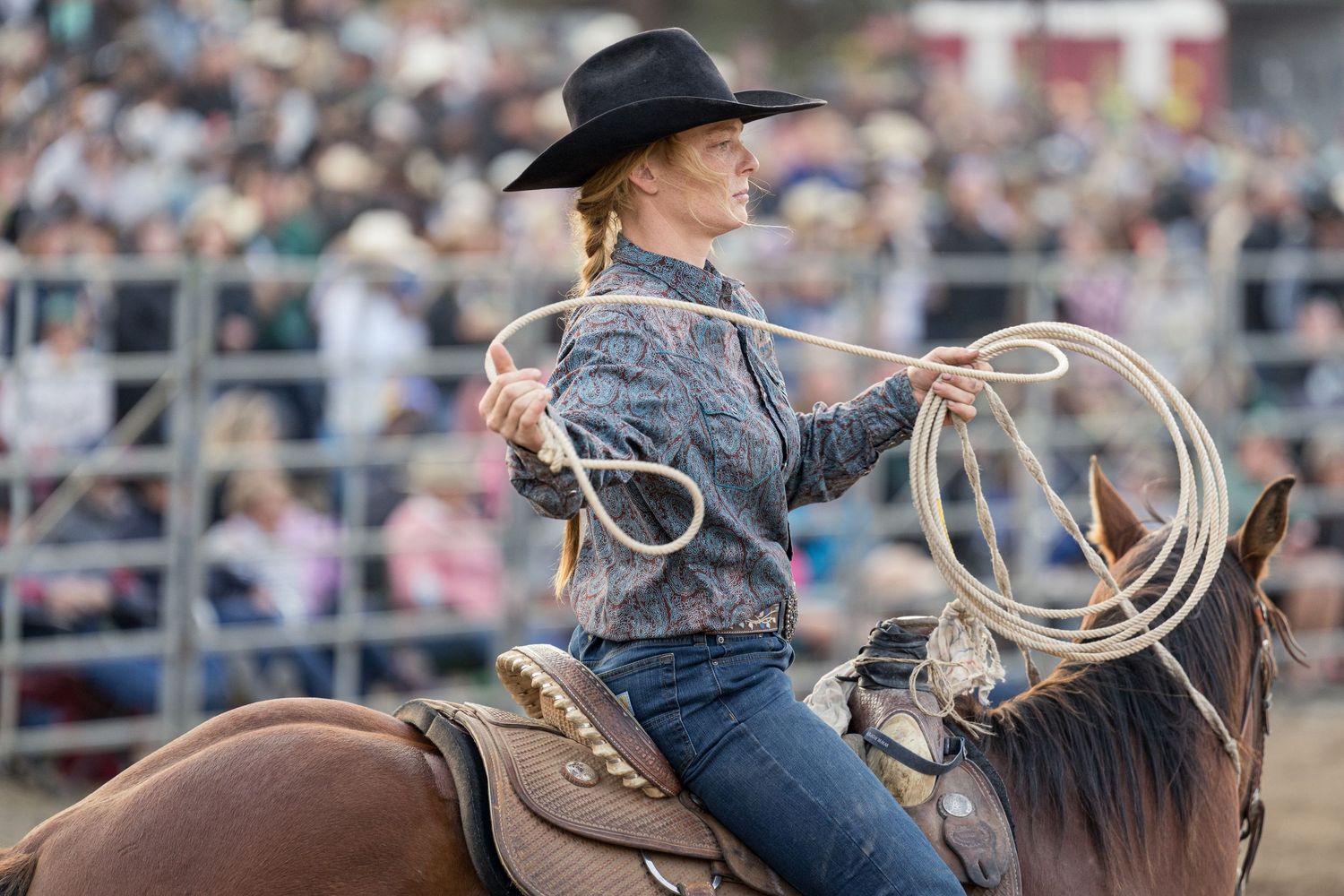 A cowgirl in a black hat rides a horse while swinging a lasso at a rodeo event.