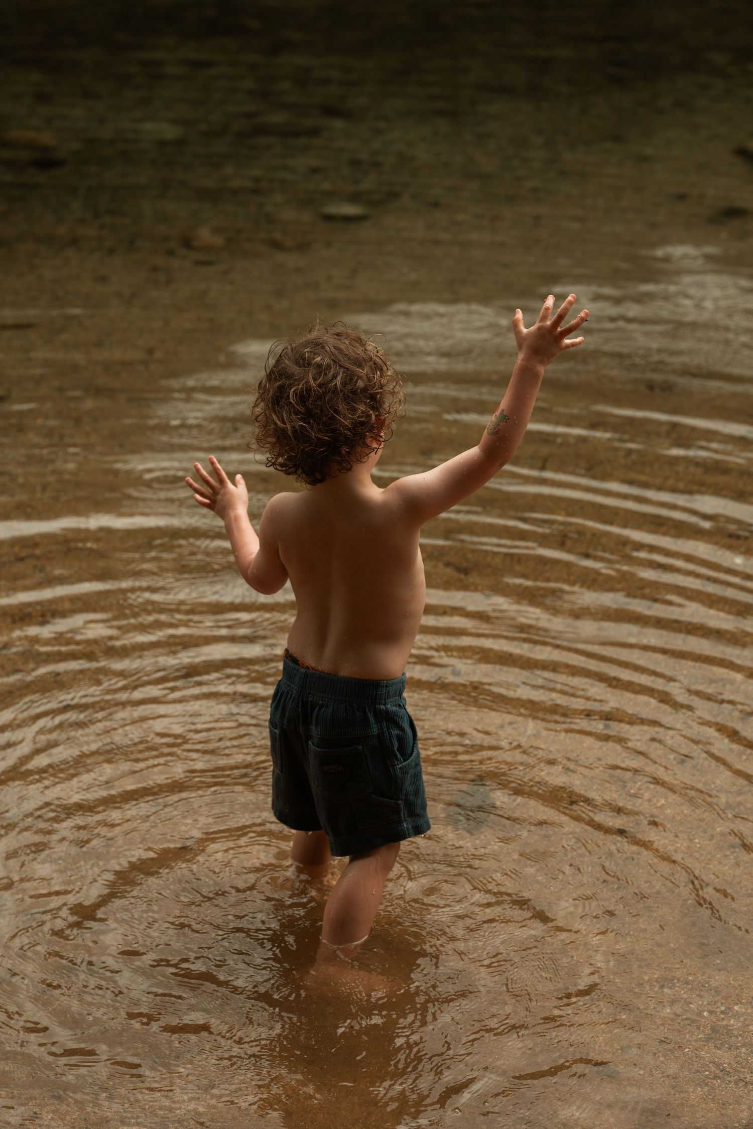 Young curly-haired child wading in shallow water with arms raised, wearing dark swim shorts.