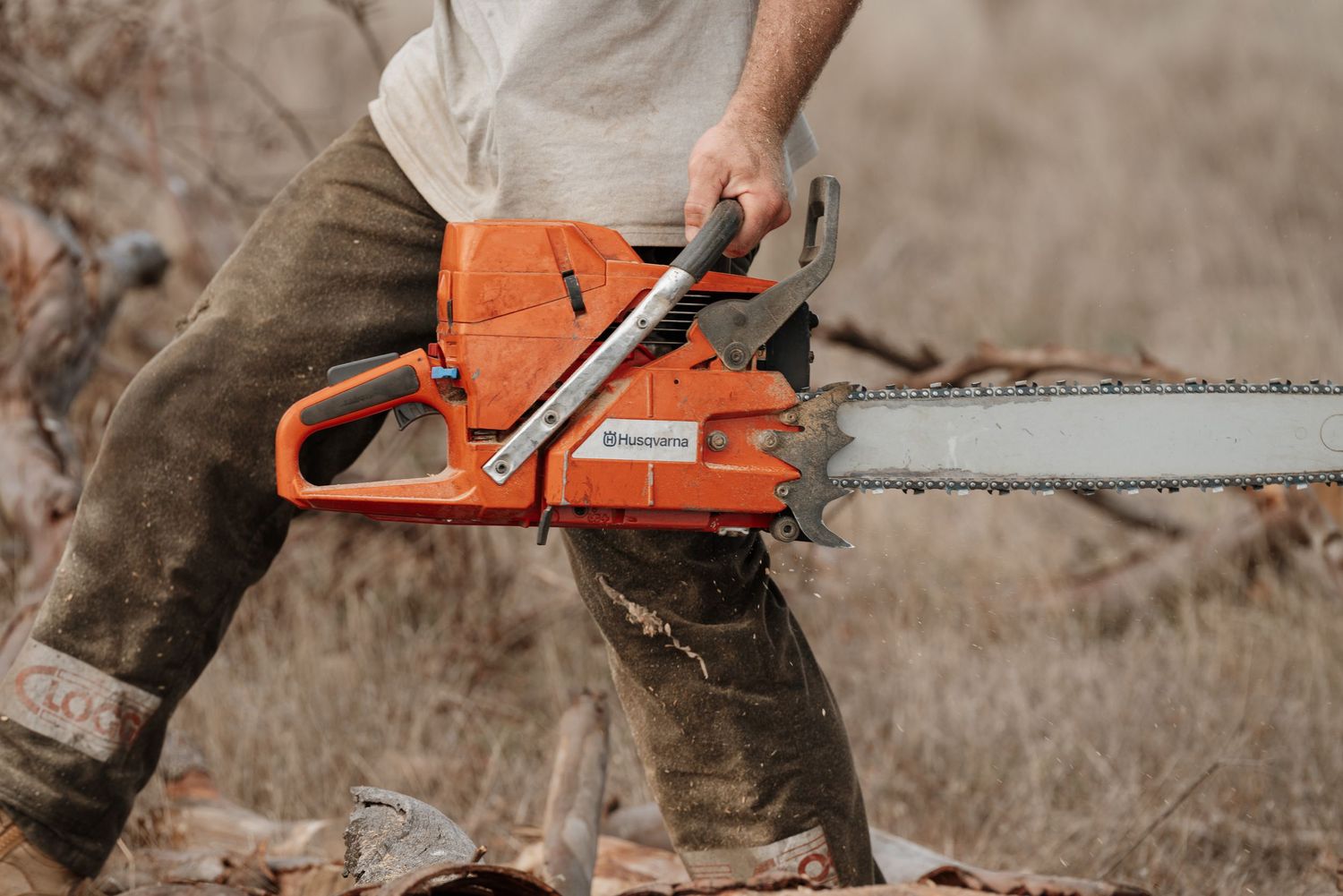 A man in dirty work clothes operating an orange Husqvarna chainsaw outdoors in a dry field.