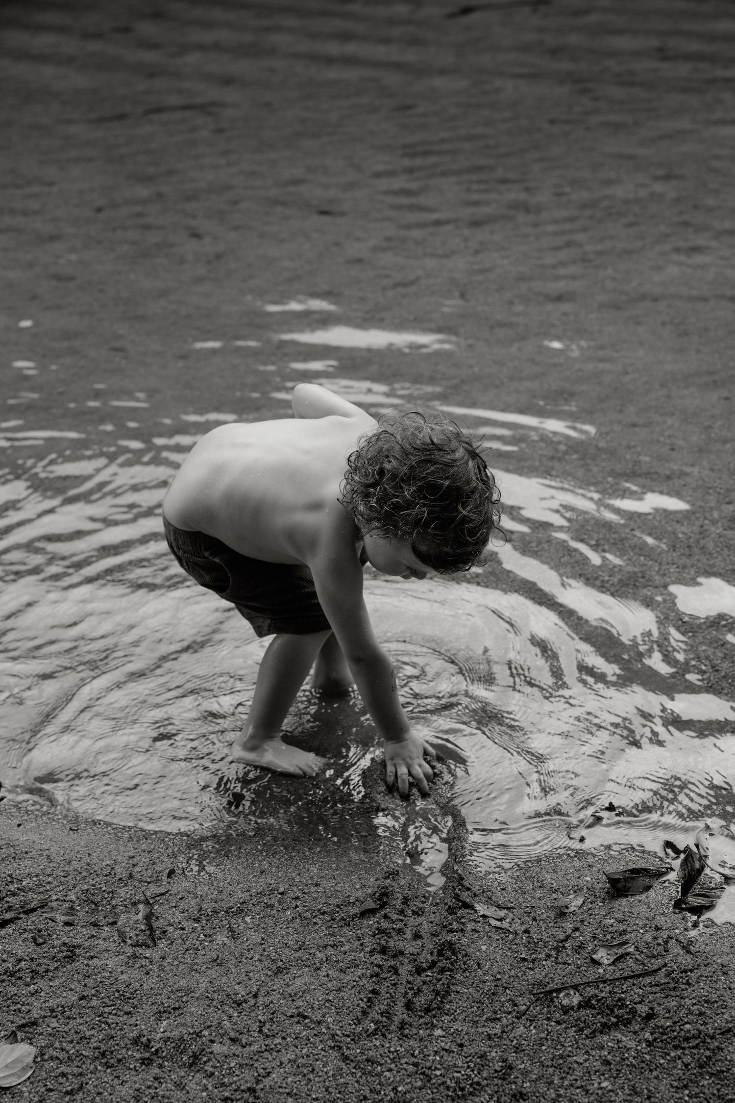 Black and white photo of a shirtless boy playing in shallow water at a sandy beach shore.
