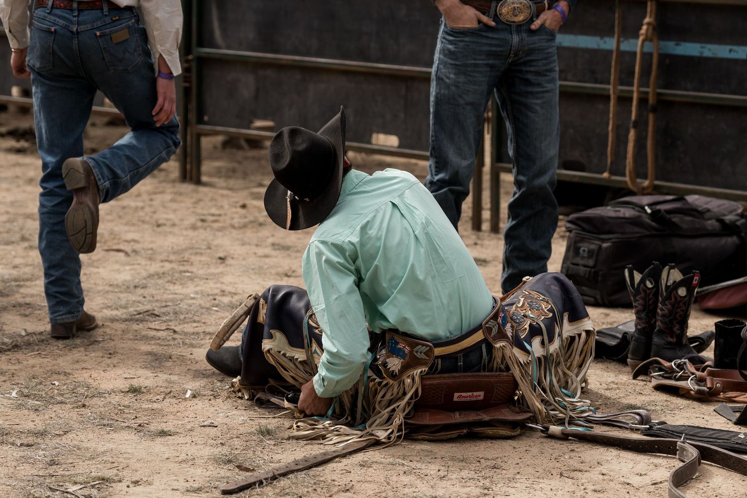 Cowboy in turquoise shirt kneeling on dirt ground adjusting leather chaps before a rodeo.