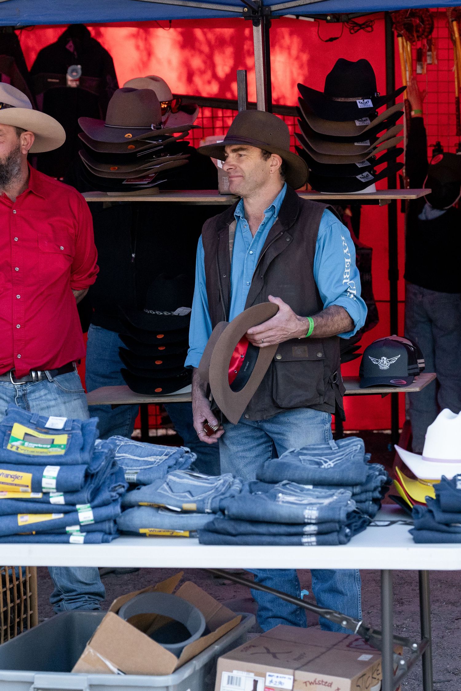 Man in cowboy hat and vest holds hat at outdoor western wear market stall with jeans and hats displayed.