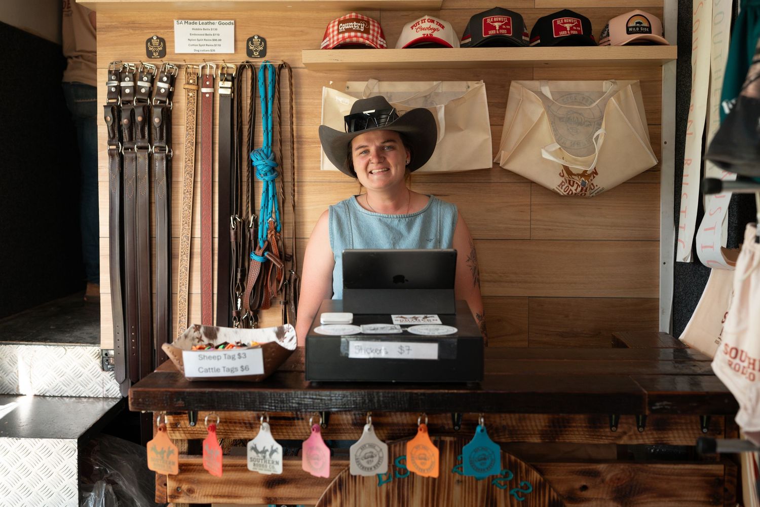 Smiling woman vendor standing at rustic wooden counter of cowboy hat and belt retail booth.