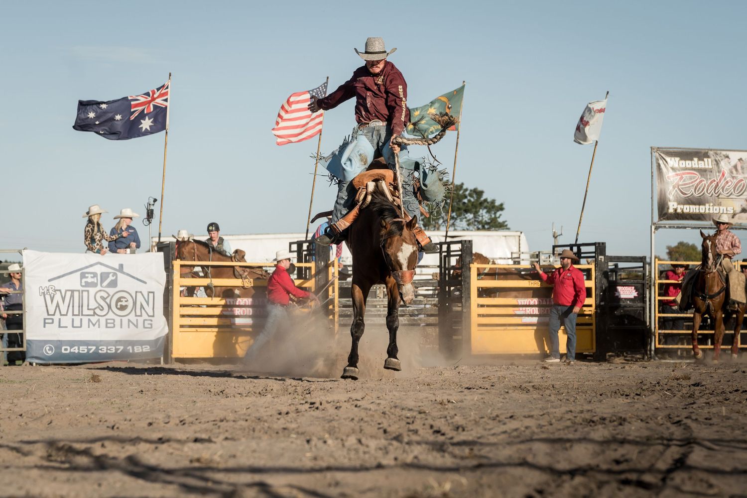 Cowboy rides bucking bronco at outdoor rodeo with Australian and American flags in background.