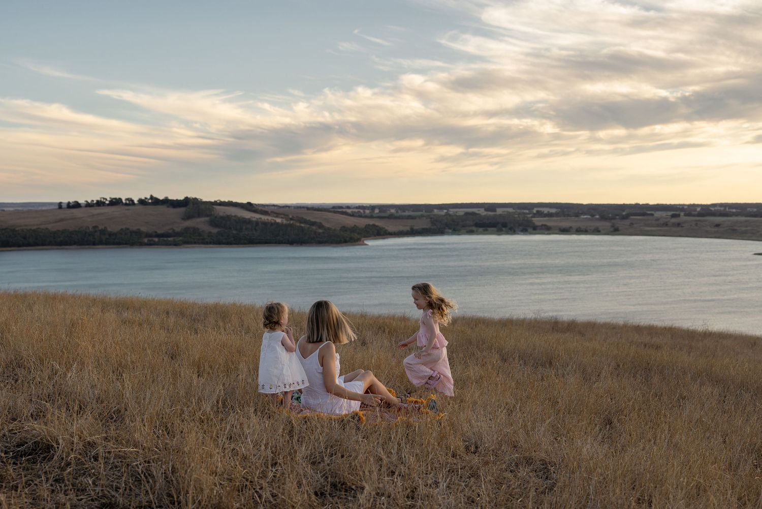 Three girls in white dresses sit in golden grass overlooking a scenic lake at sunset.
