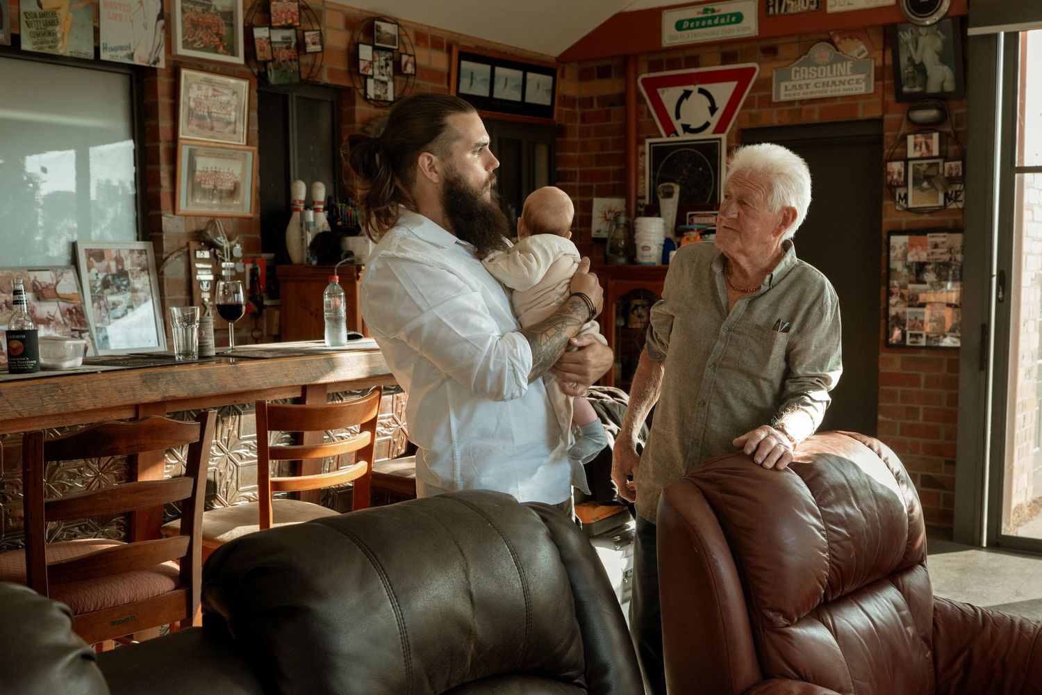 Bearded man holding baby while elderly man sits in leather chair in a cozy room.