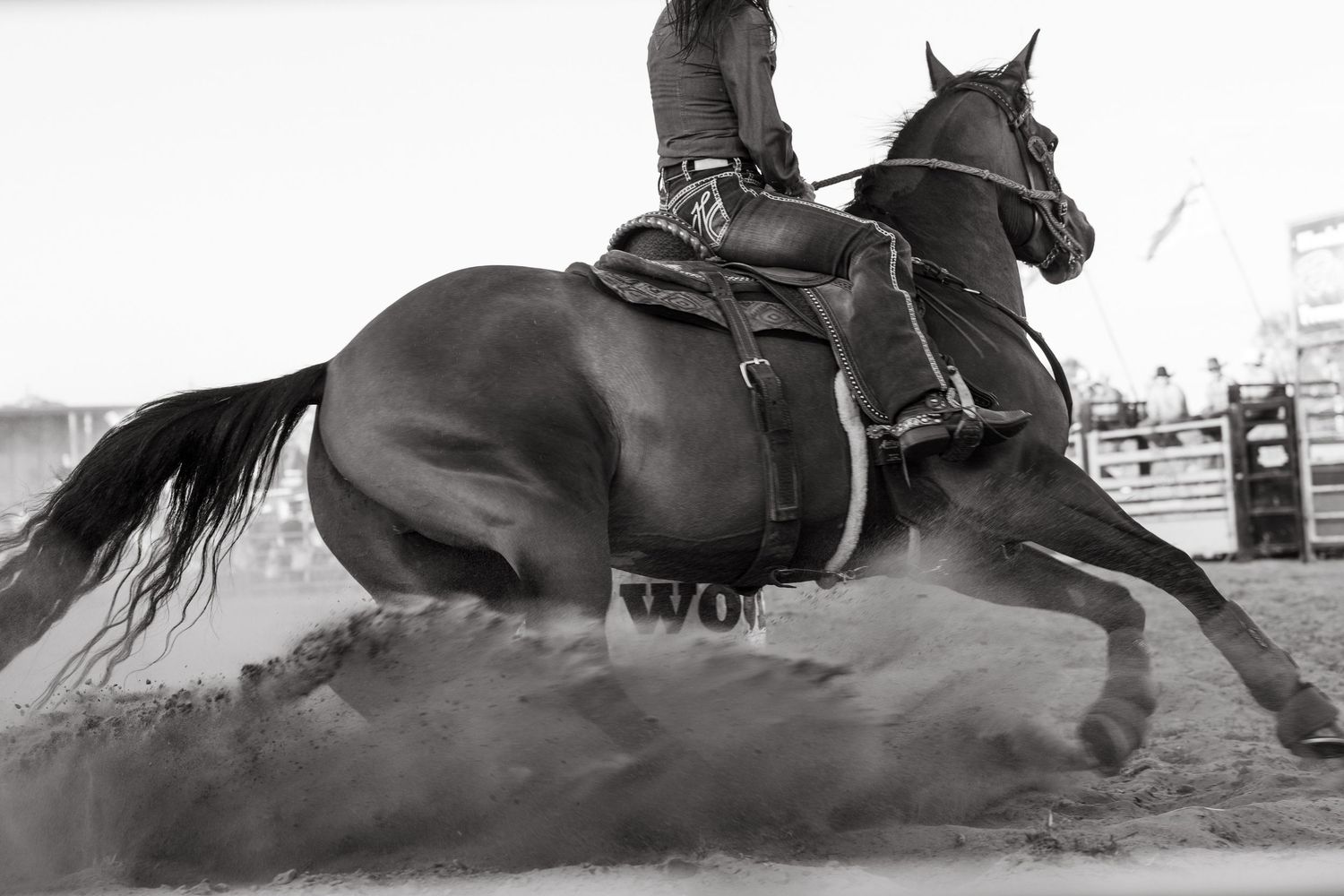 Black and white photo of a cowgirl on a dark horse barrel racing in a dusty rodeo arena.