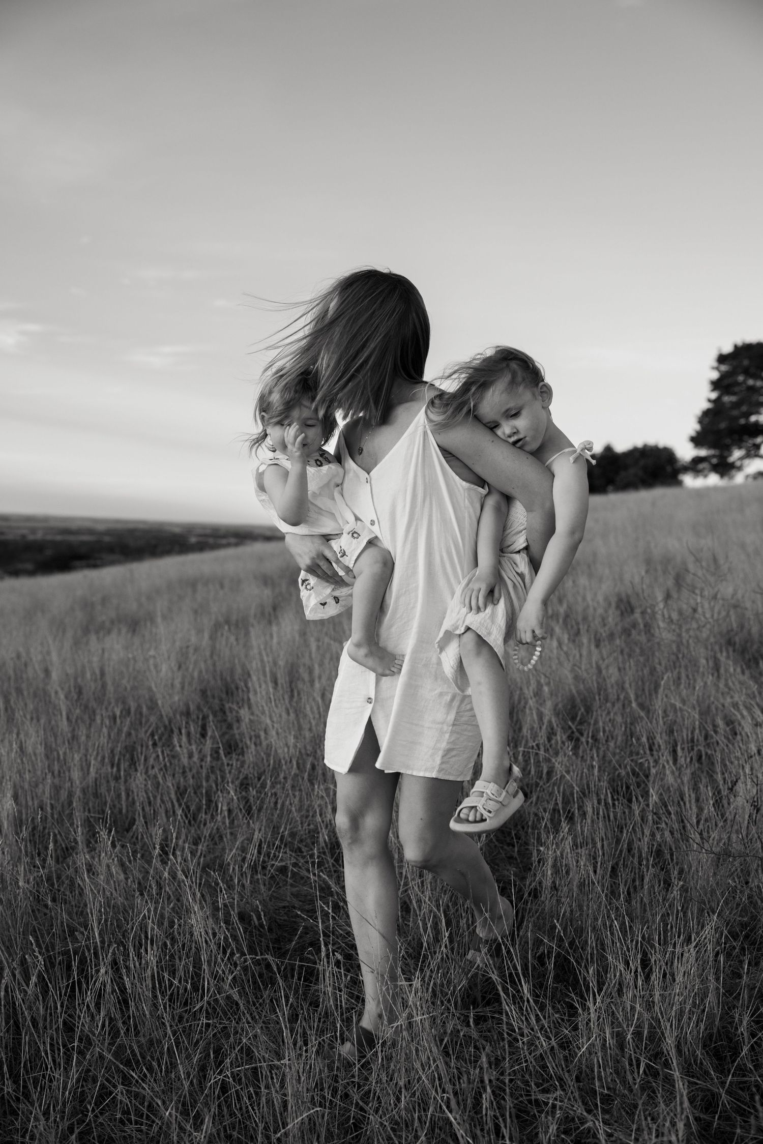 Black and white photo of a mother carrying two young children through a grassy field at sunset.