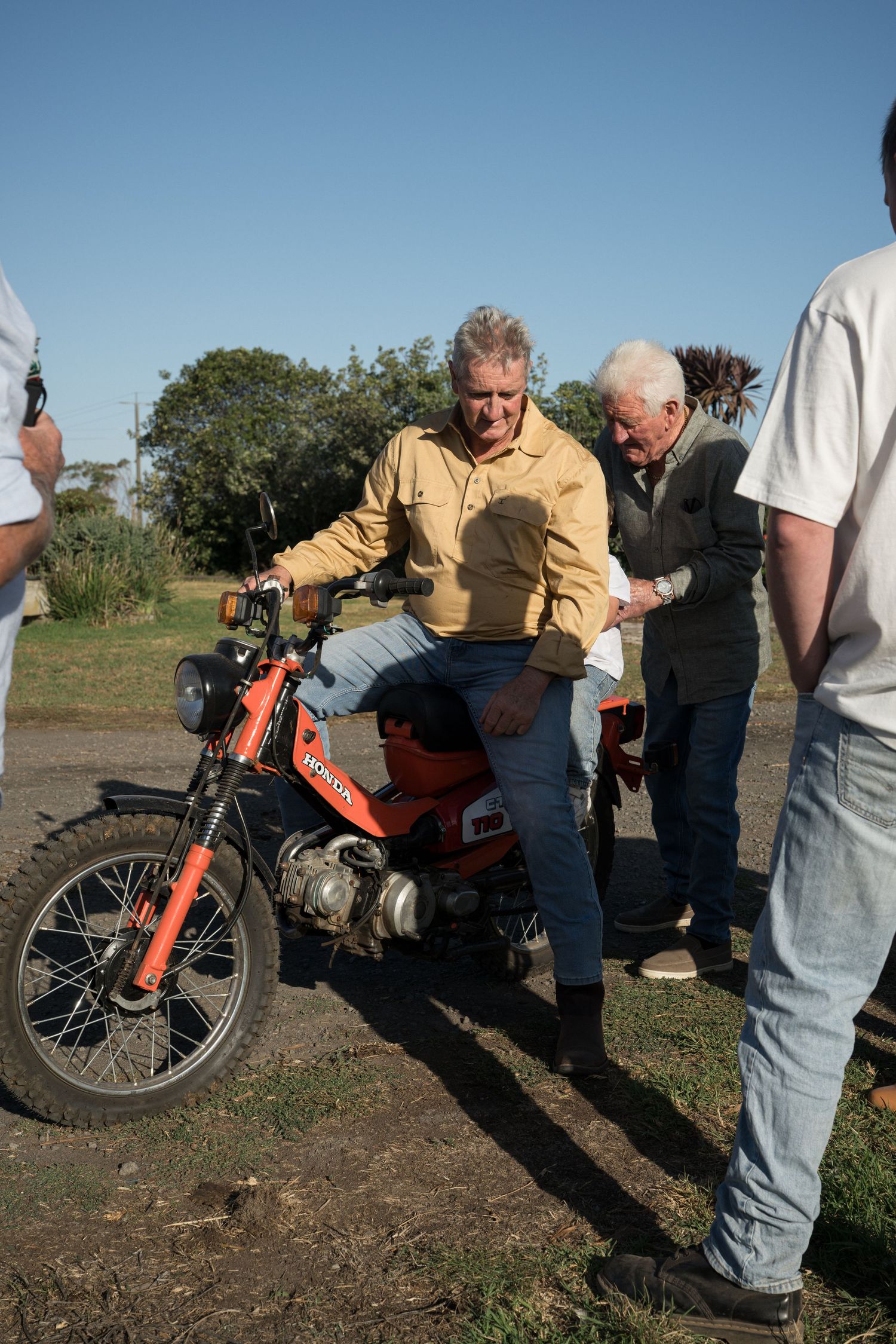 Men examining a vintage orange Honda motorcycle outdoors on a sunny day.