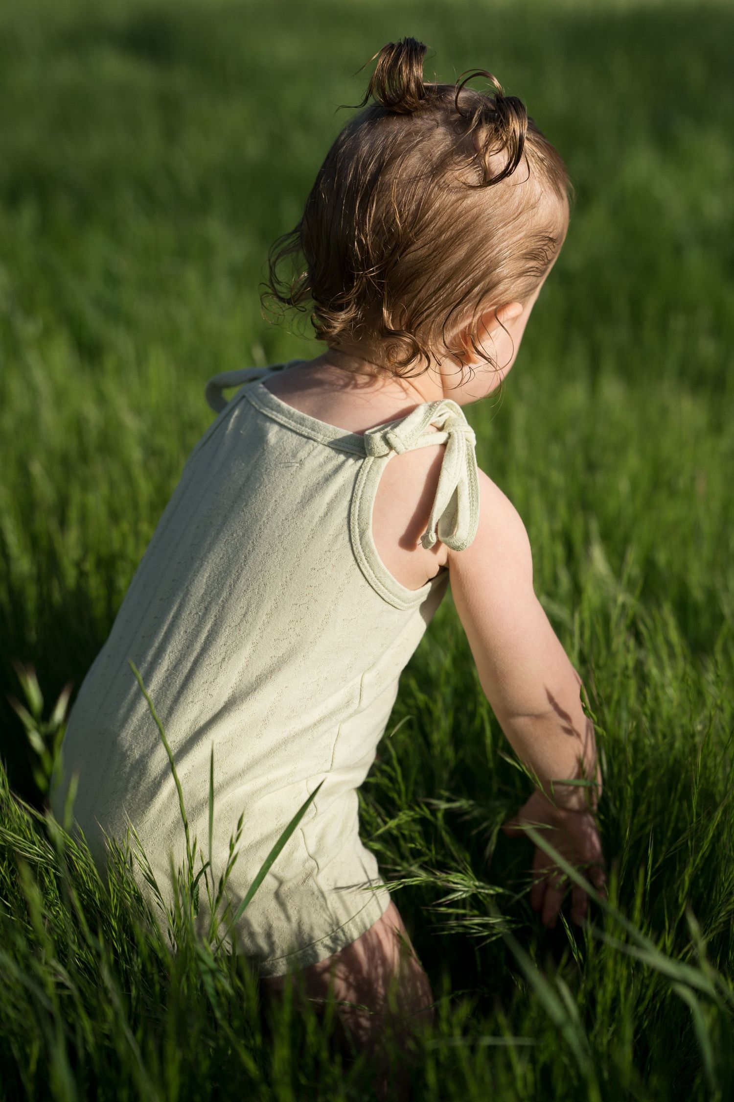 Toddler girl in light green romper kneels in tall green grass in warm sunlight.