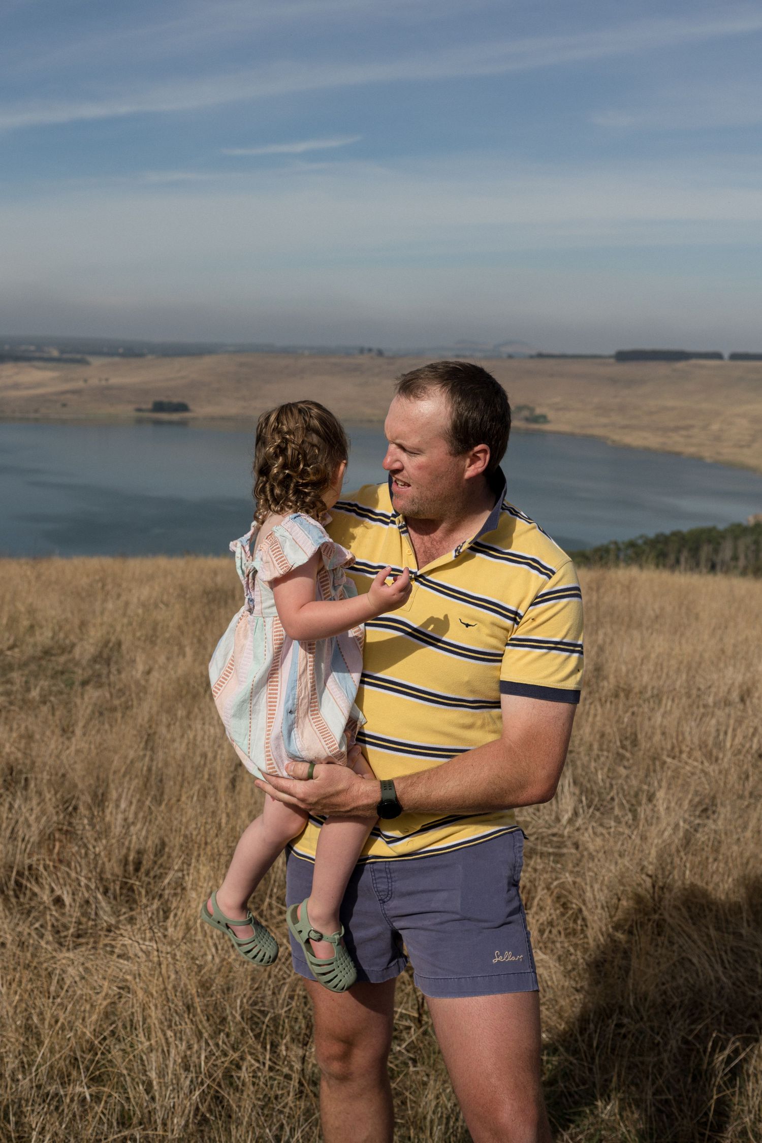 Father in yellow striped polo shirt holding young daughter in open grassland with lake backdrop.