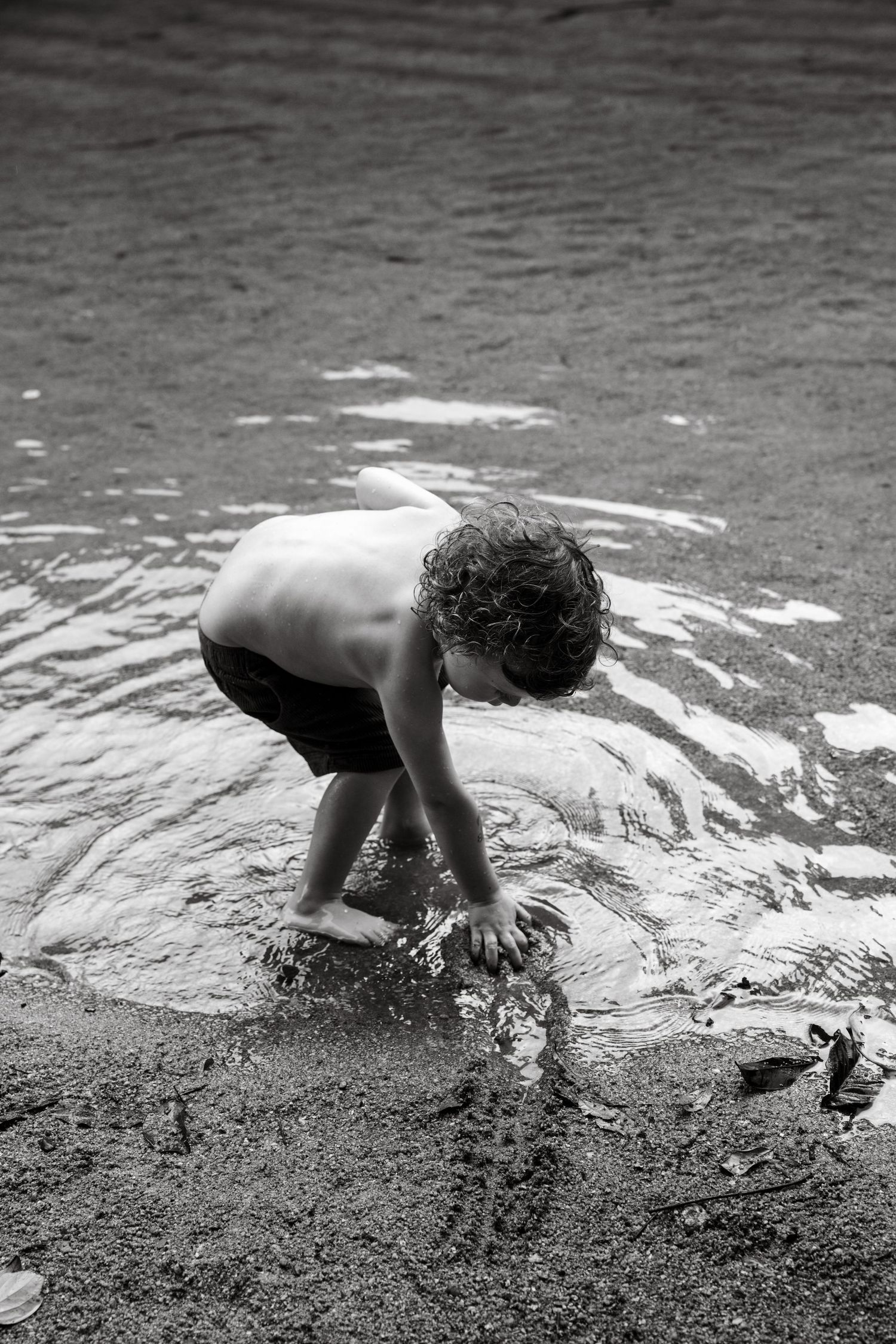 Black and white photo of a child playing in shallow water on a sandy river shore.