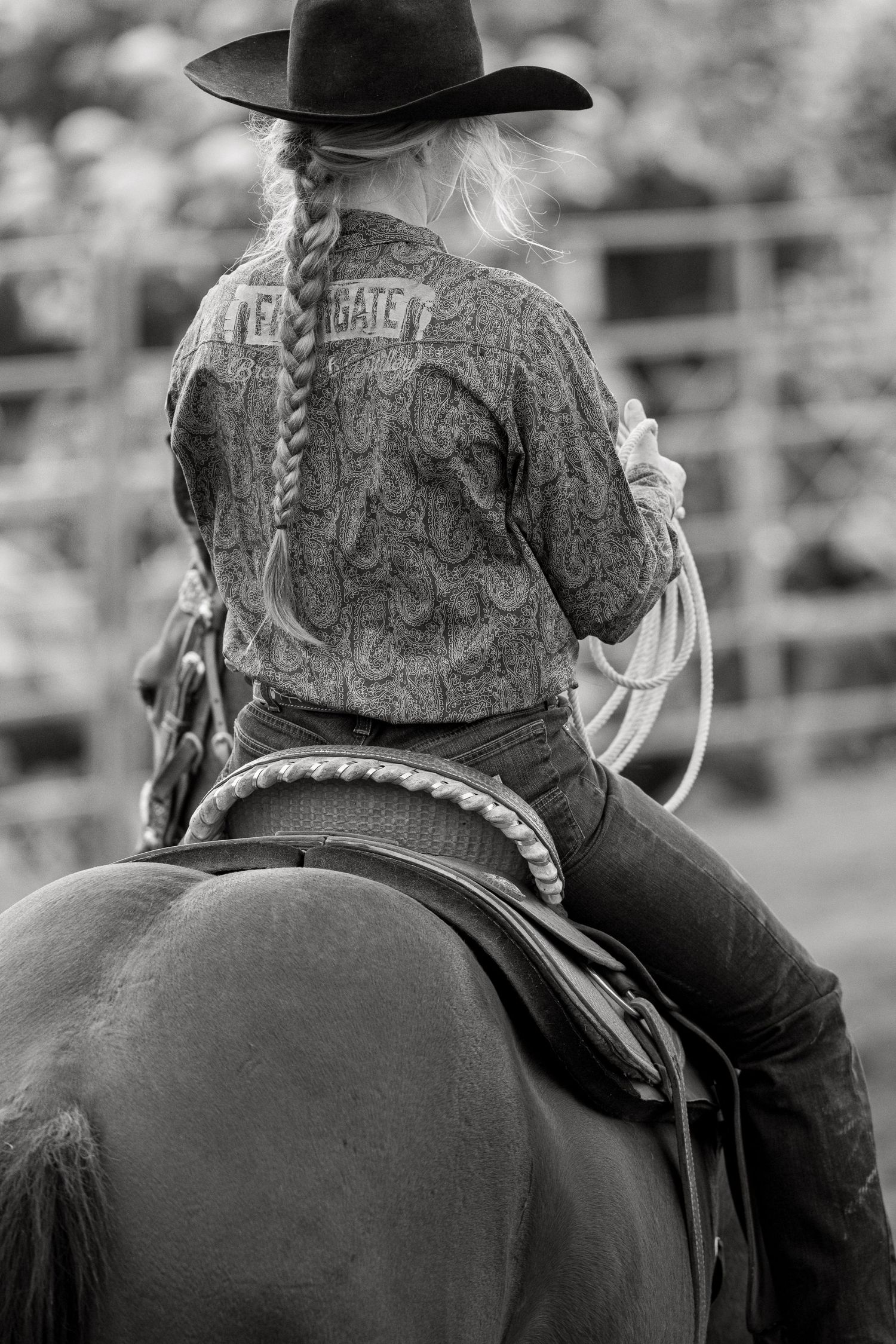 Black and white rear view of a cowgirl on horseback holding a rope at a rodeo.
