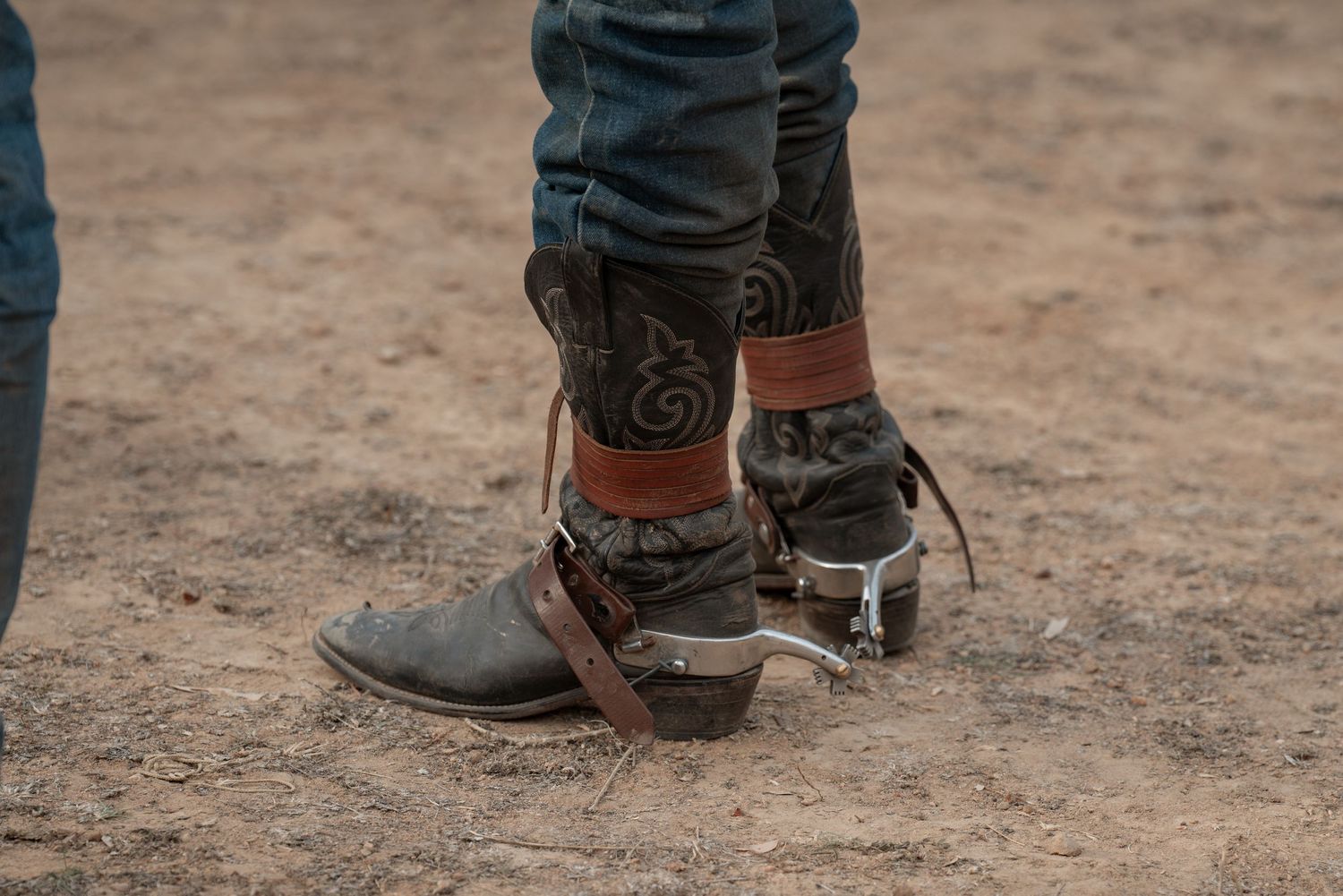 Close-up of cowboy boots with decorative stitching and leather spurs standing on dirt ground.