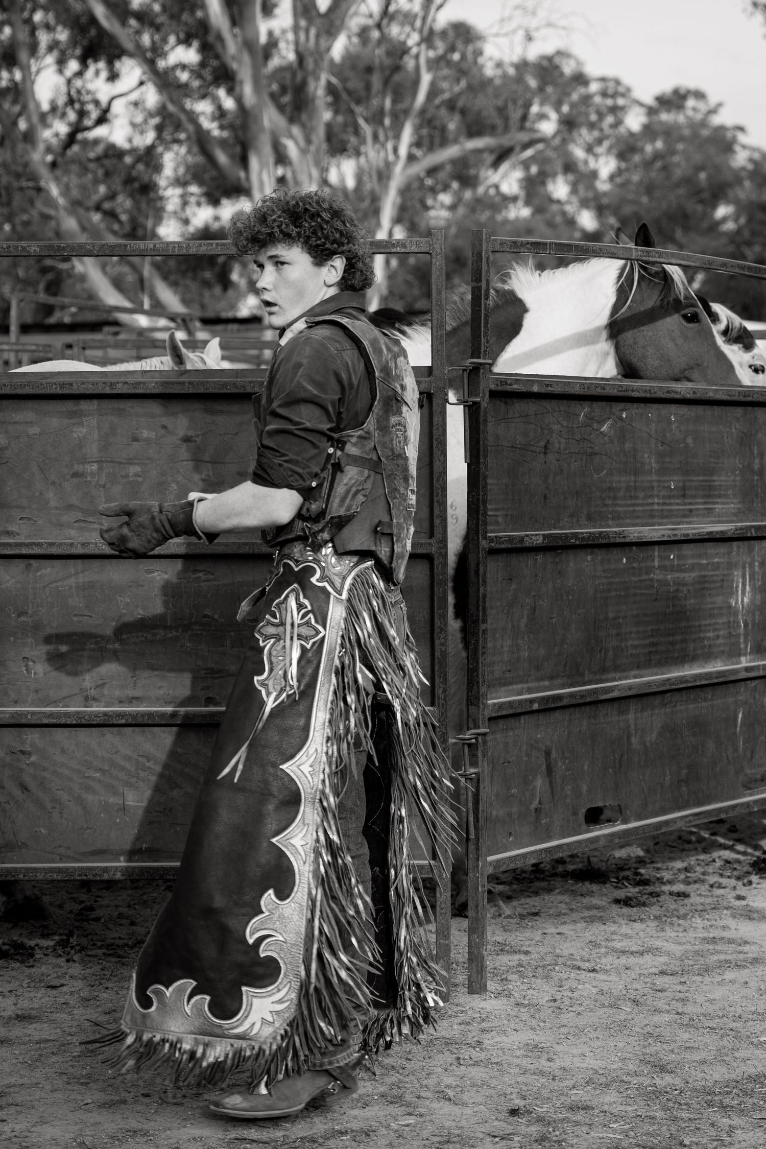 Black and white photo of a young cowboy in chaps standing by a rodeo gate.
