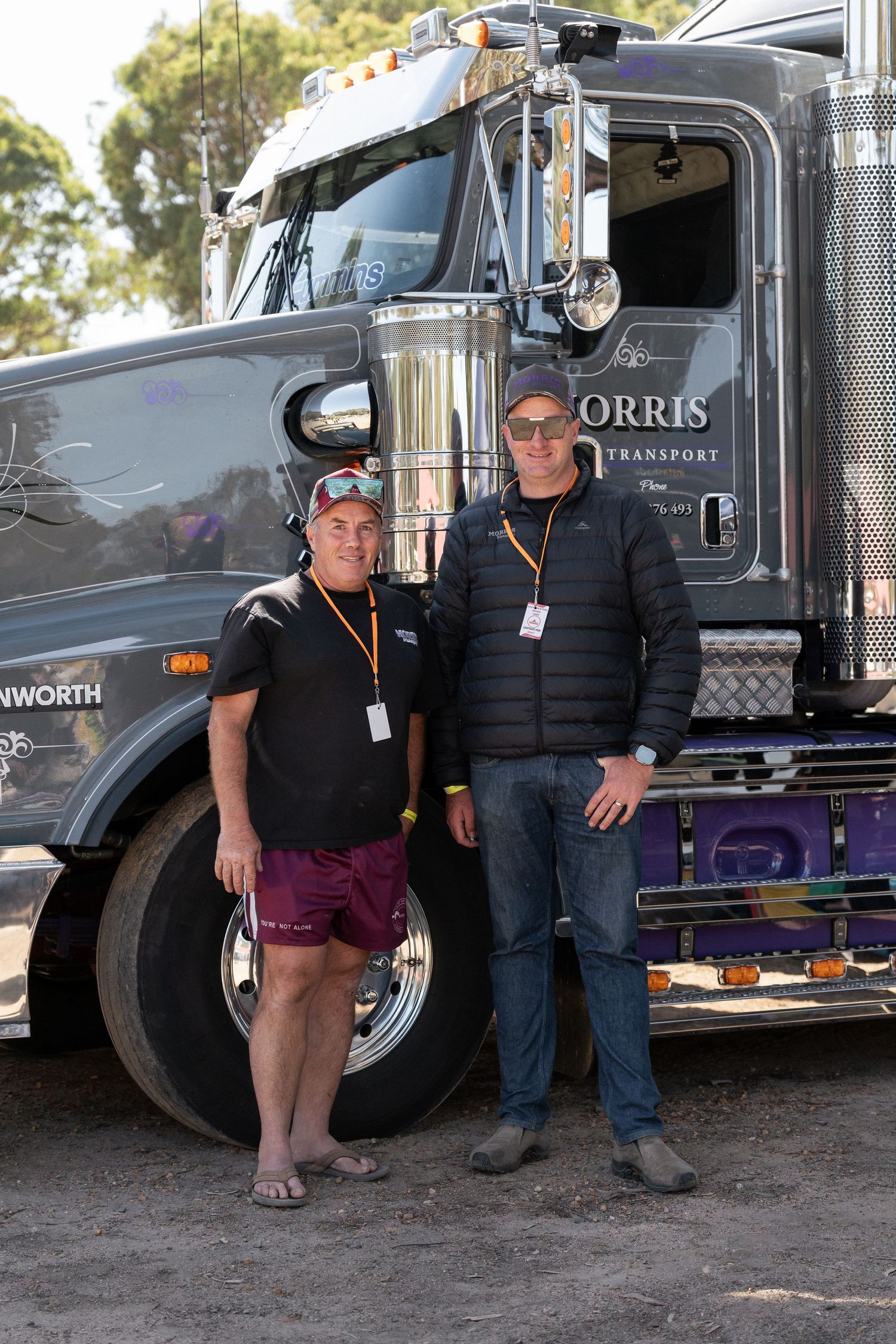 Two men smiling and posing in front of large Kenworth Morris Transport semi-truck outdoors.