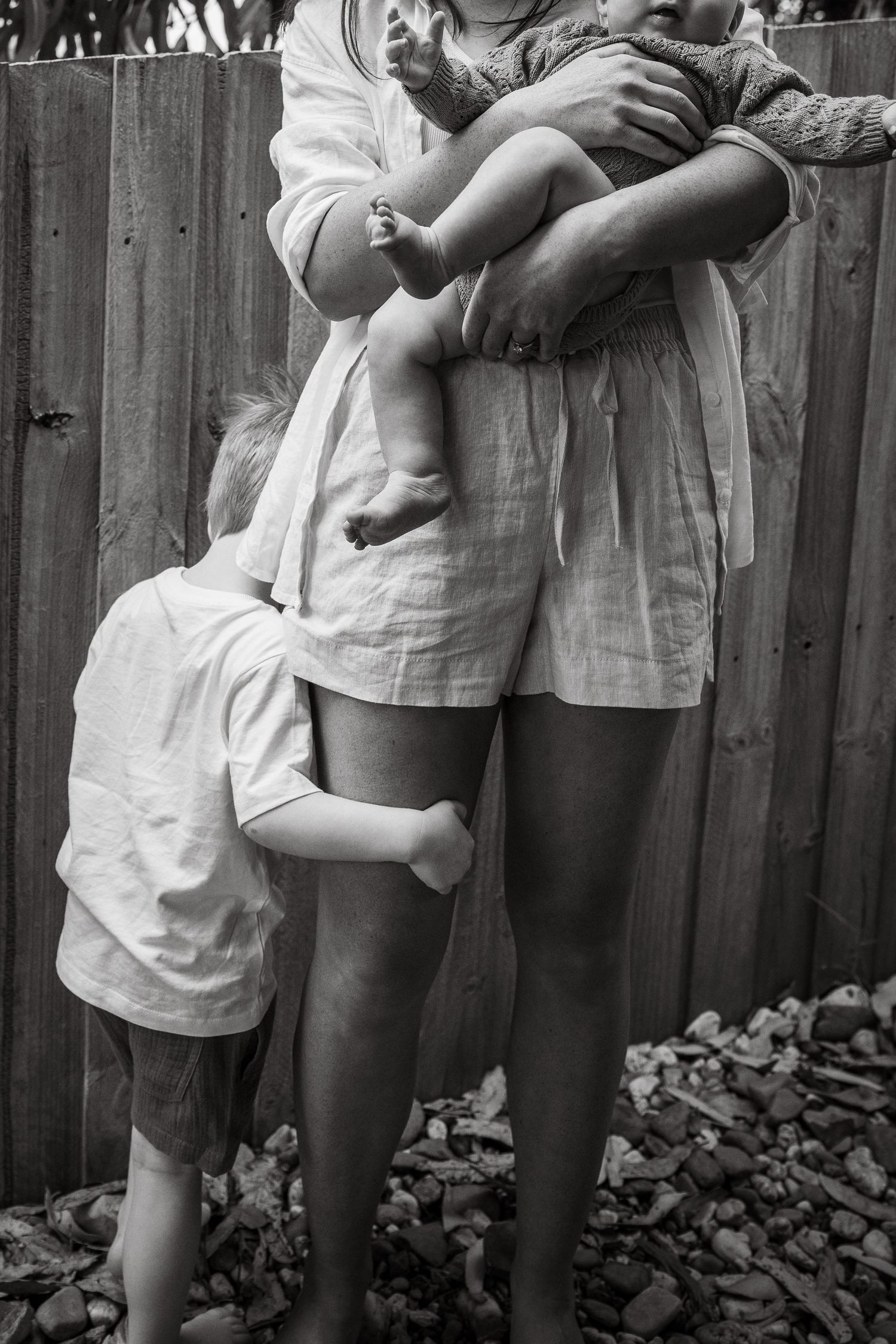 Black and white photo of a mother holding a baby while a young child hugs her legs near a wooden fence.