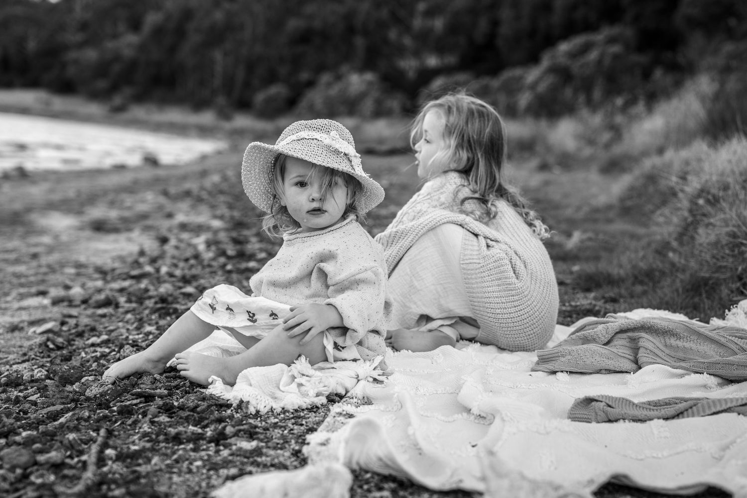 Two young children sit on a blanket at a rocky beach, one wearing a sun hat, in a black and white photo.