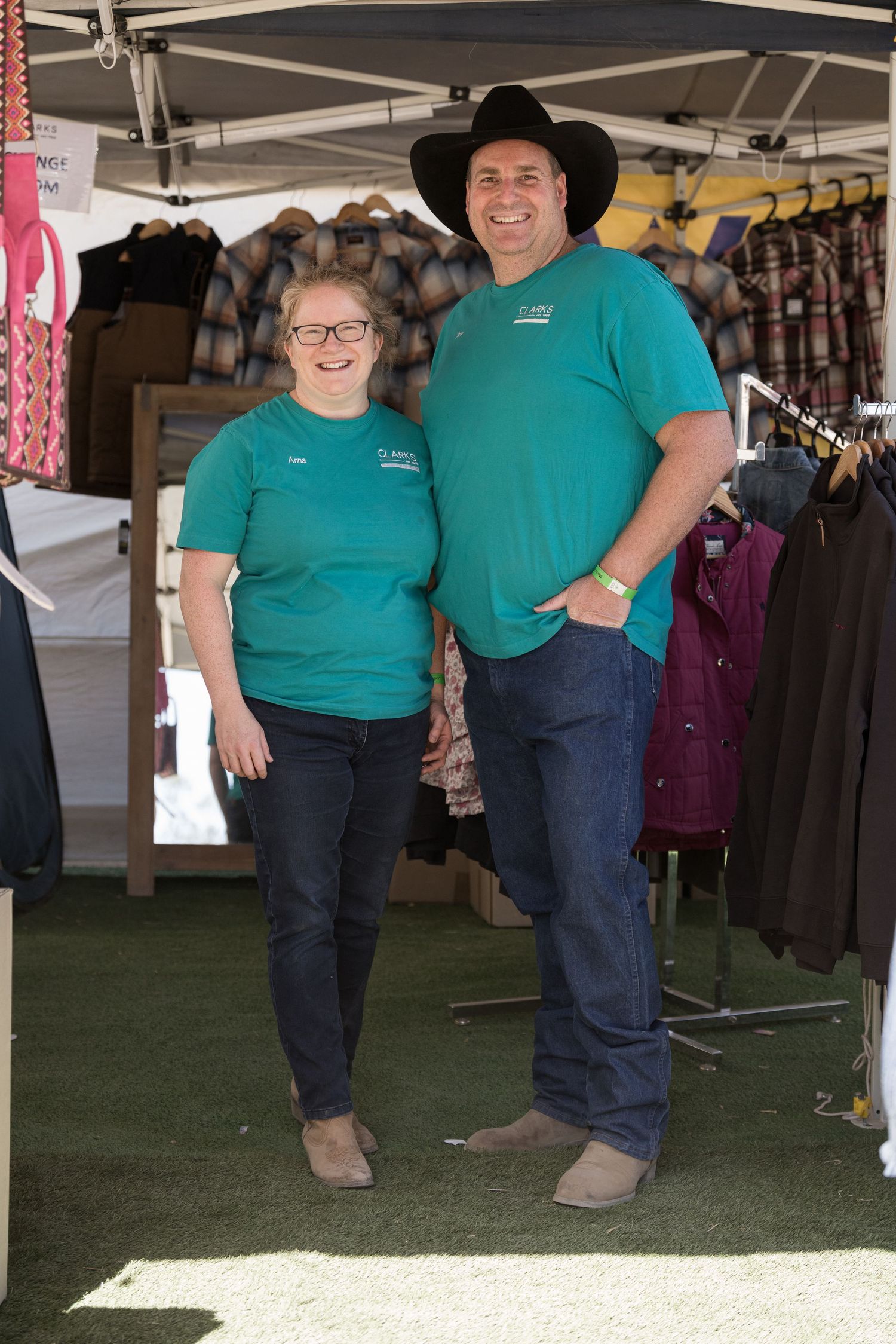 Smiling couple in matching teal shirts pose together at outdoor western clothing market stall.