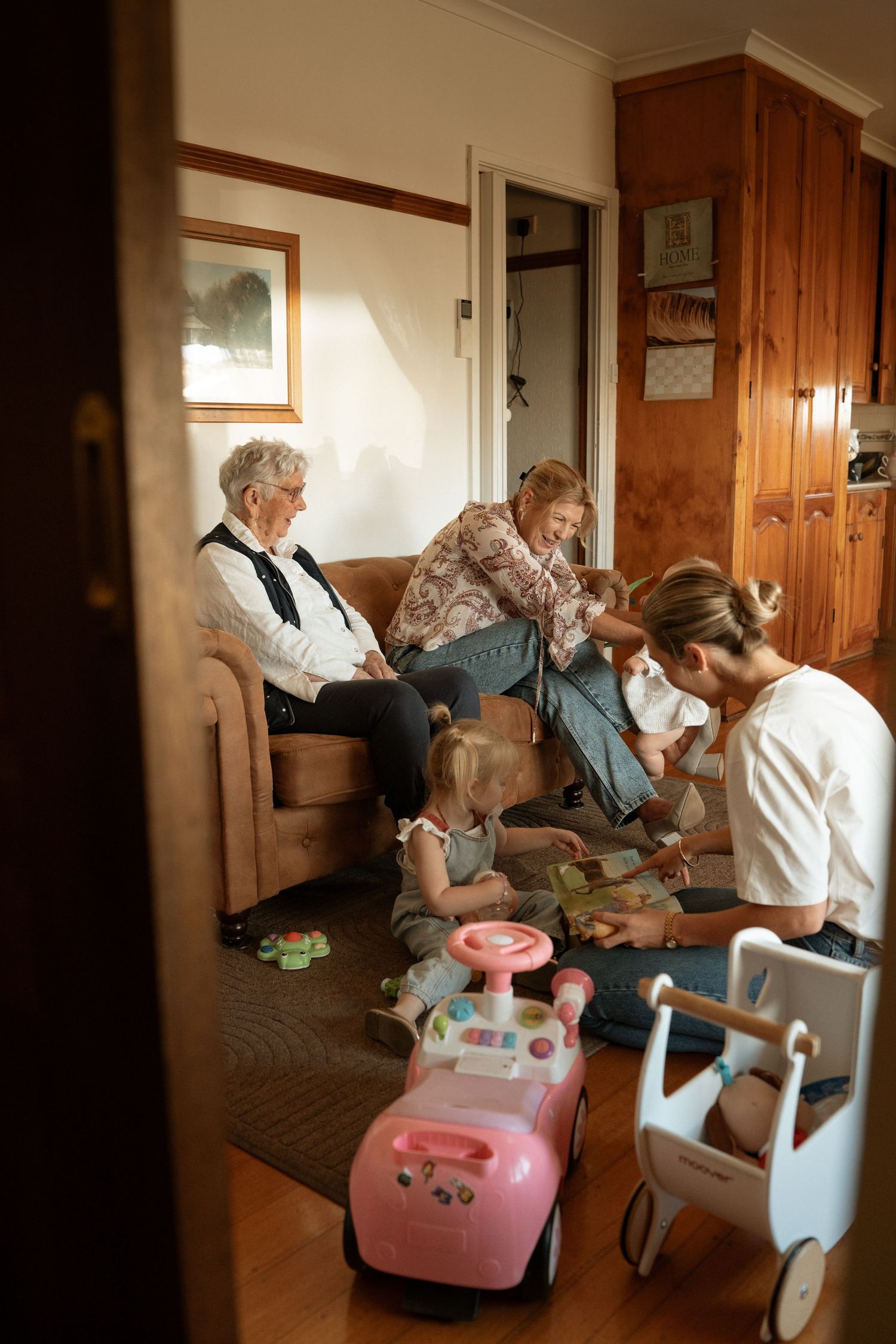 Multi-generational family gathering indoors with grandmother, mother, and children playing with toys on living room floor.