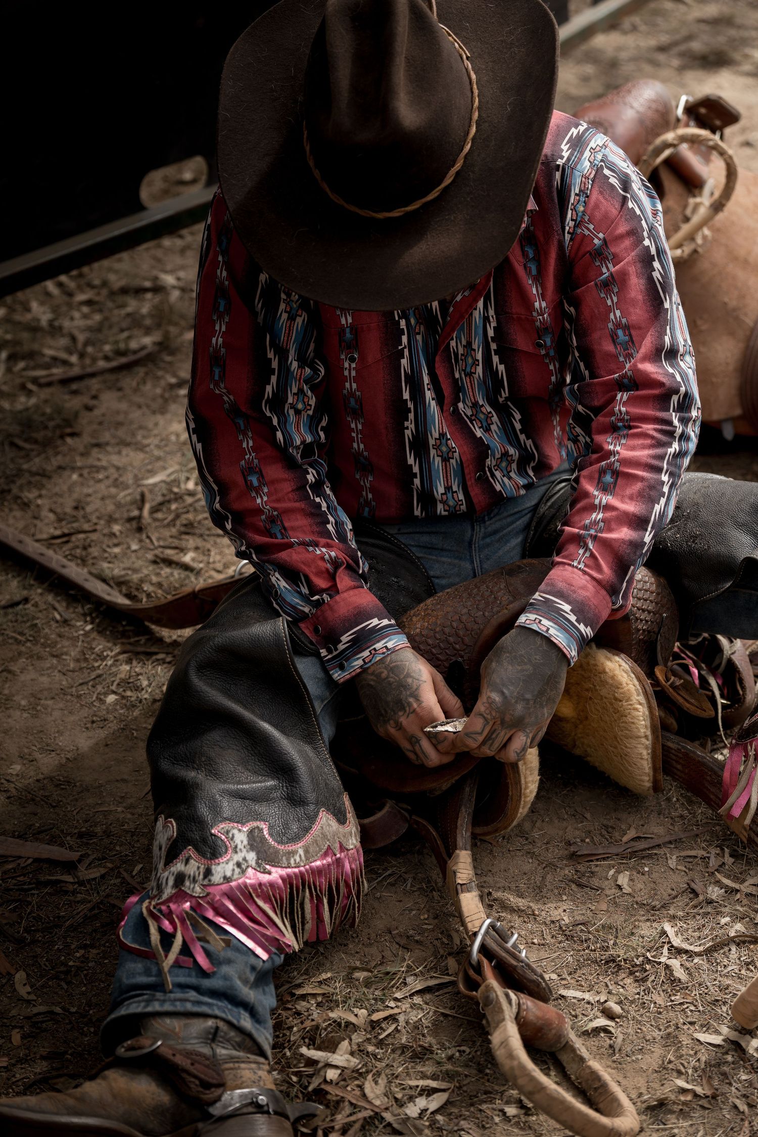 Cowboy in decorative western shirt and chaps sits on ground adjusting spurs at rodeo.
