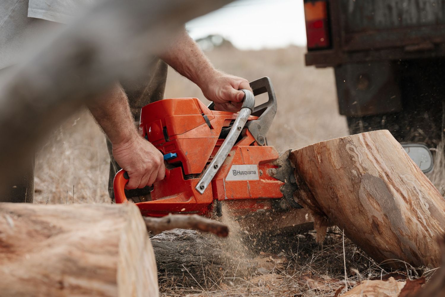 Man using an orange Husqvarna chainsaw to cut large logs outdoors near a truck.