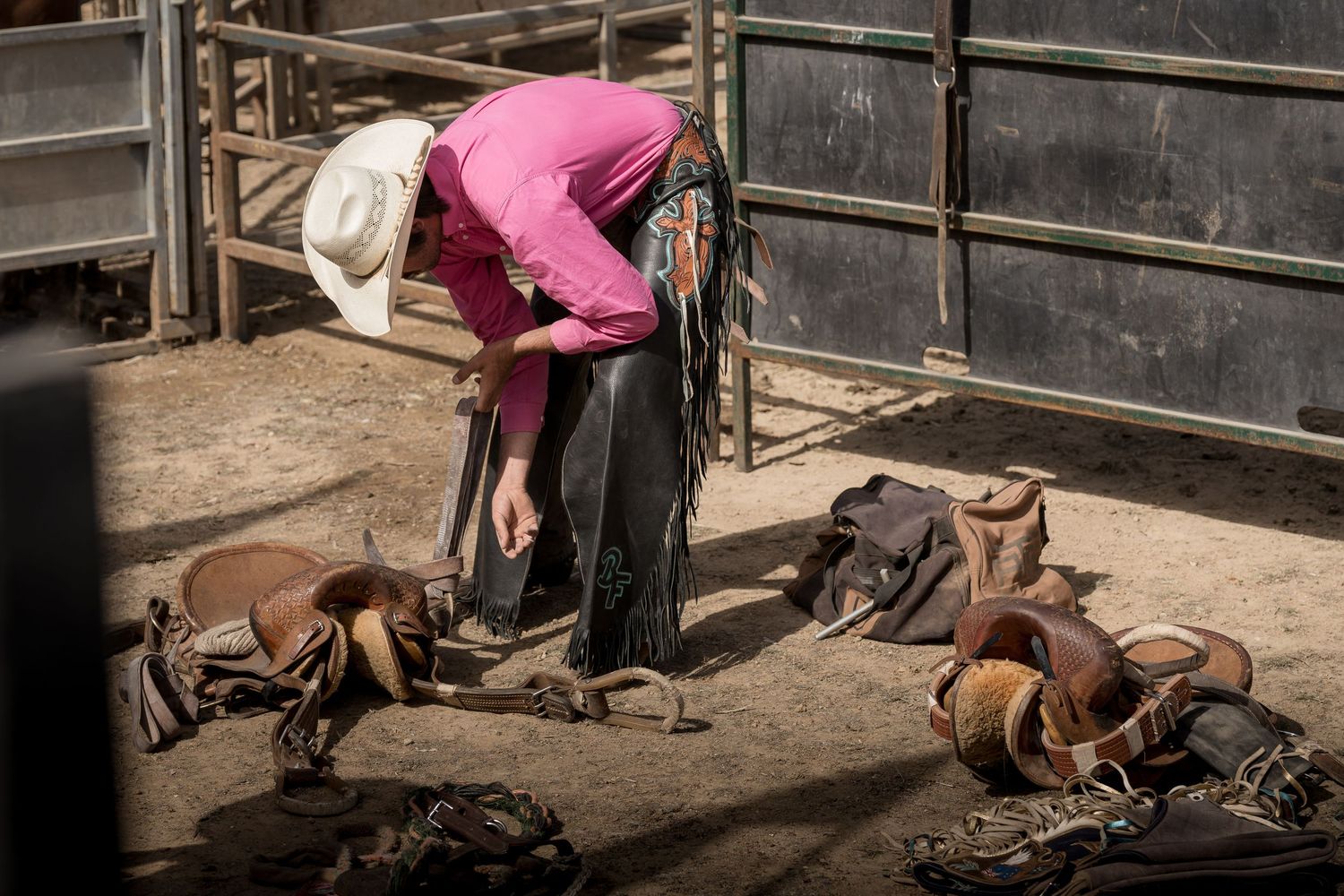 Cowboy in pink shirt bending over to adjust leather chaps surrounded by rodeo equipment.