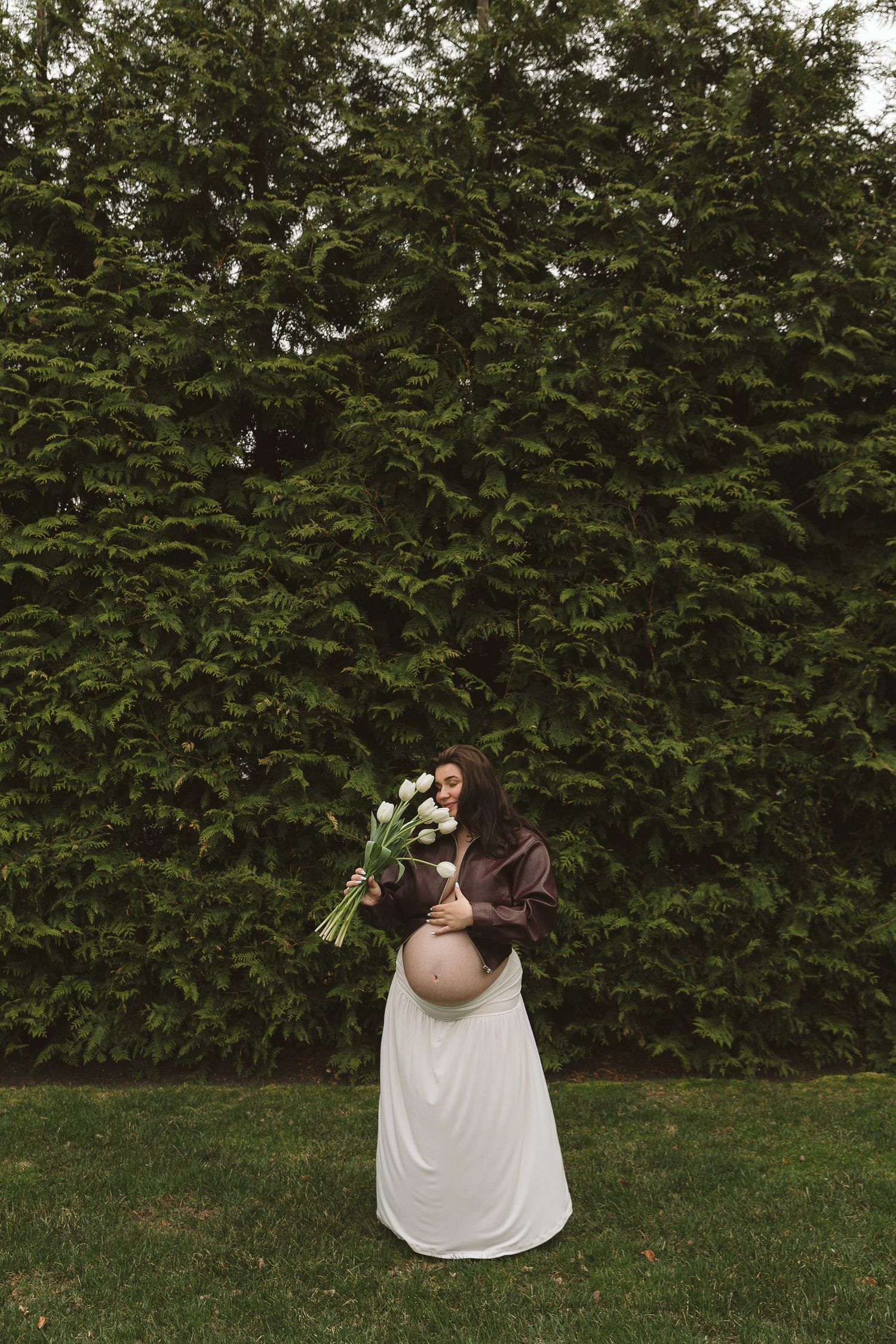 Pregnant woman in white skirt and brown top holds white tulips bouquet outdoors against lush green hedge wall.