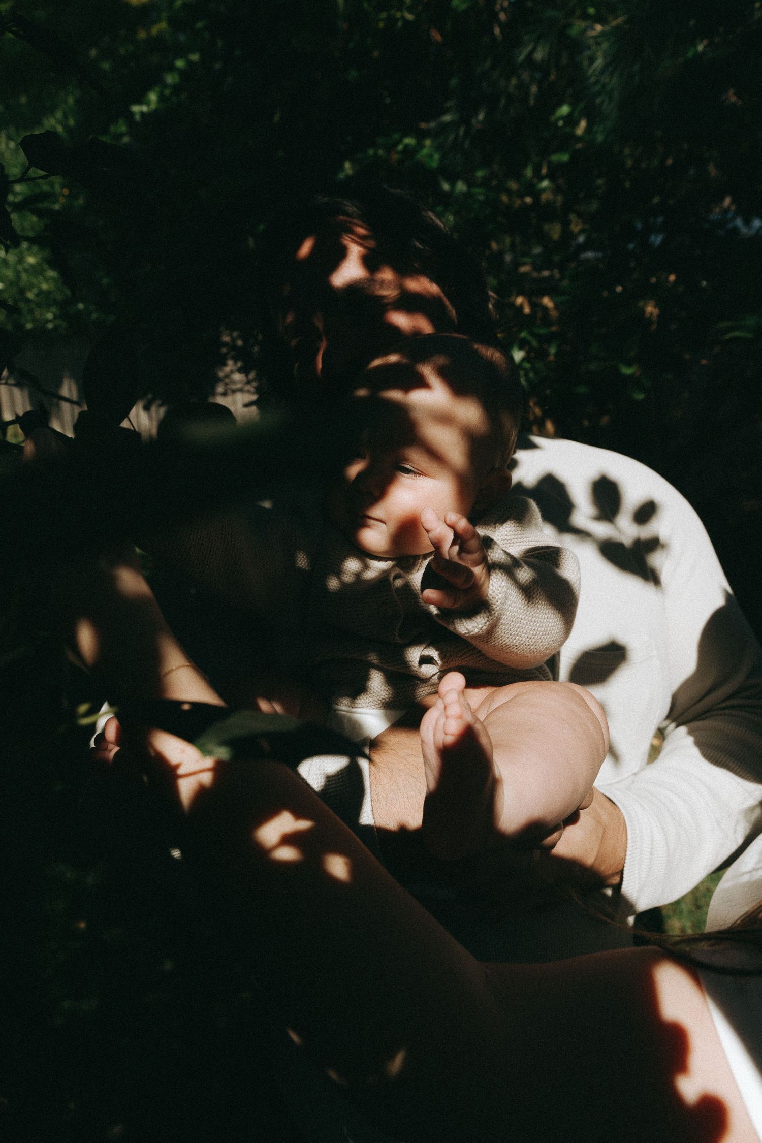 A person sits outdoors in dappled sunlight with leaf shadows cast across their face and white shirt.