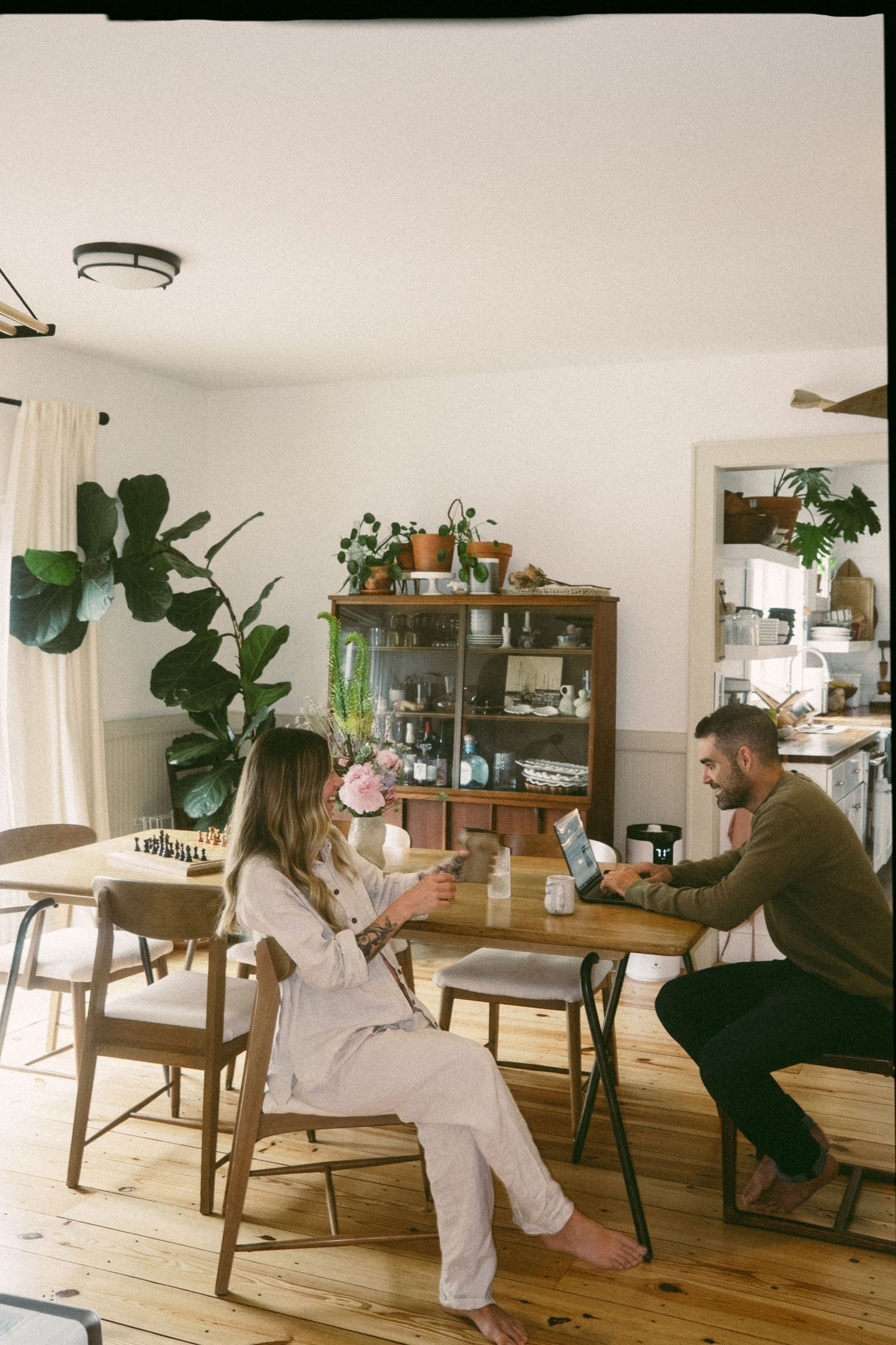 A person in white overalls lounges in a bright living room with wooden furniture and plants.