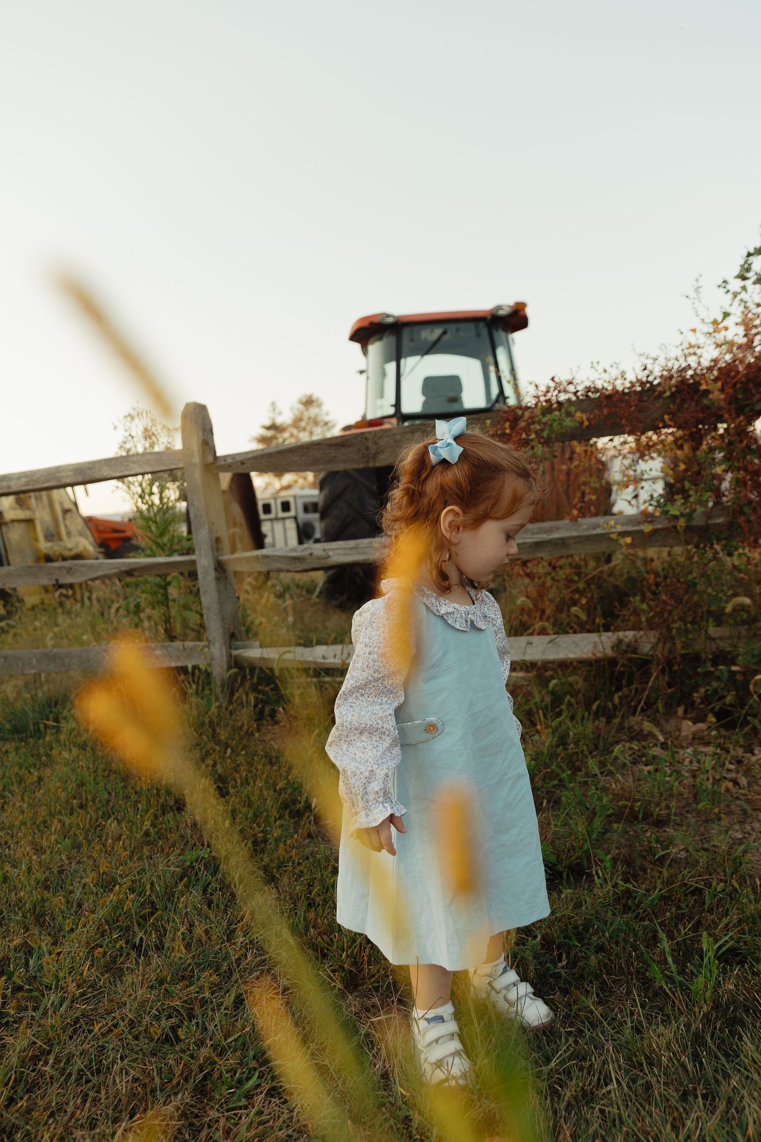 A young red-haired girl in a light blue dress stands by a wooden fence near a red farm tractor.