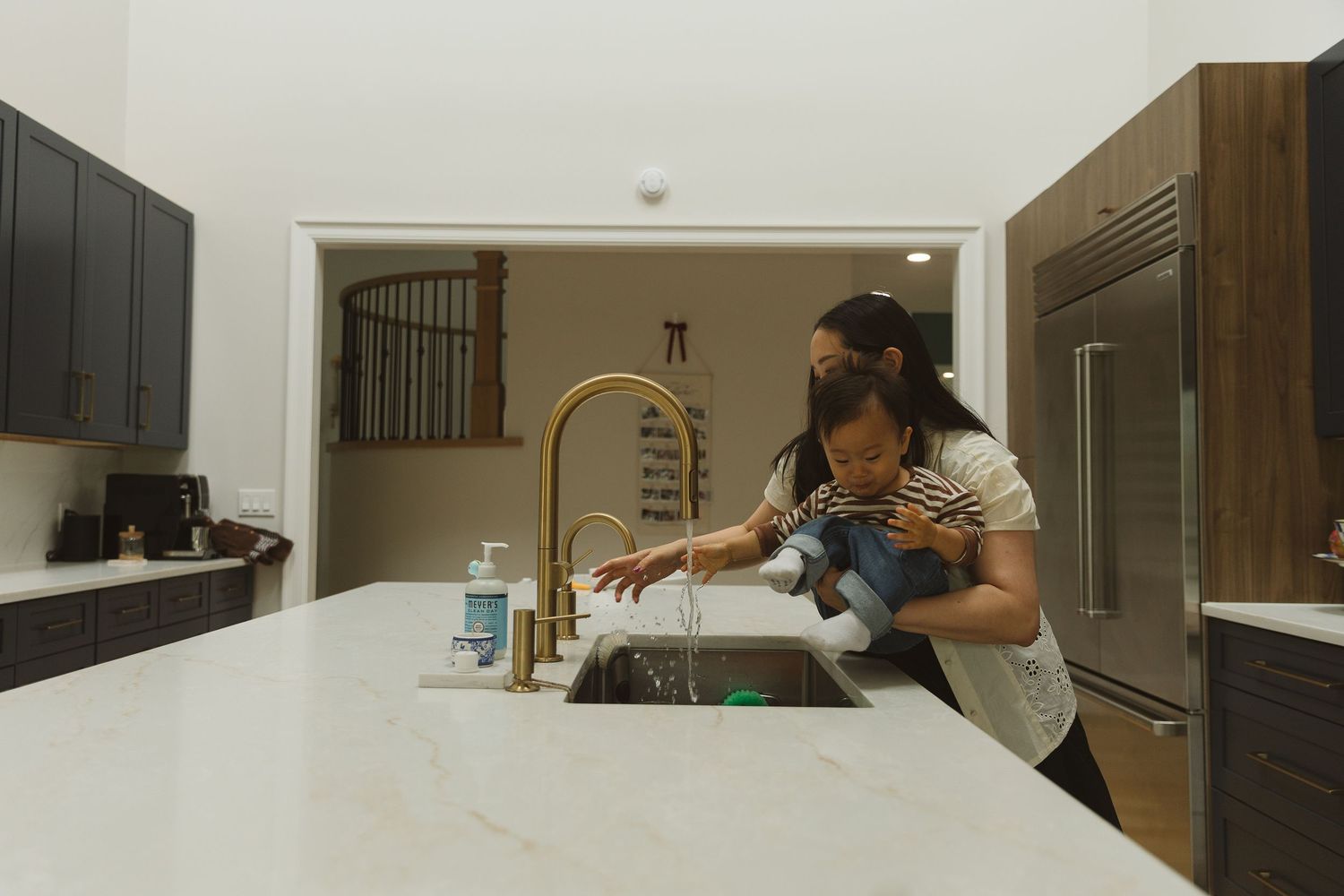 Mother washing toddler's hands at a gold kitchen faucet on a white marble island in a modern home.