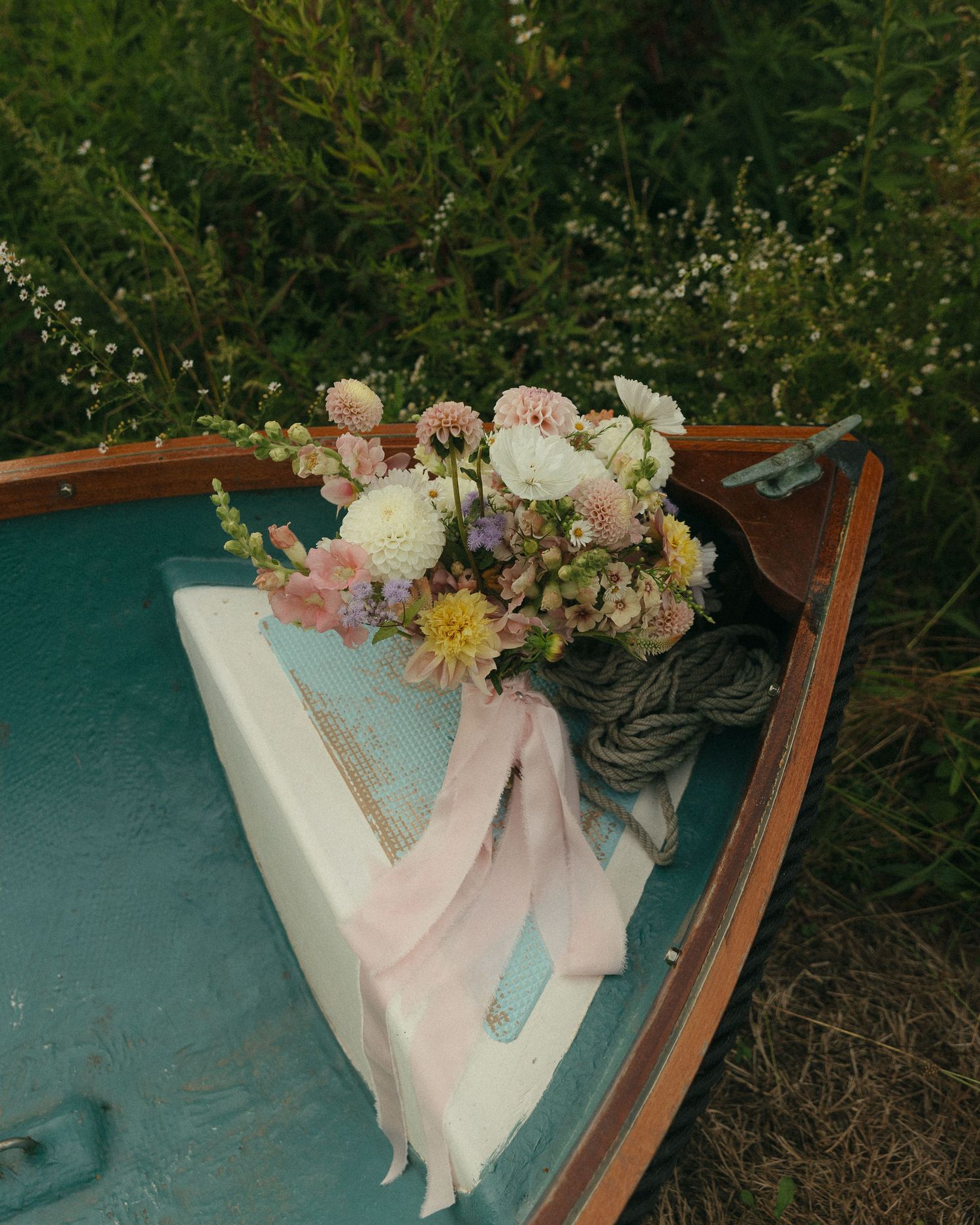 A rustic floral bouquet with pink and white blooms rests on a teal wooden boat surrounded by greenery.