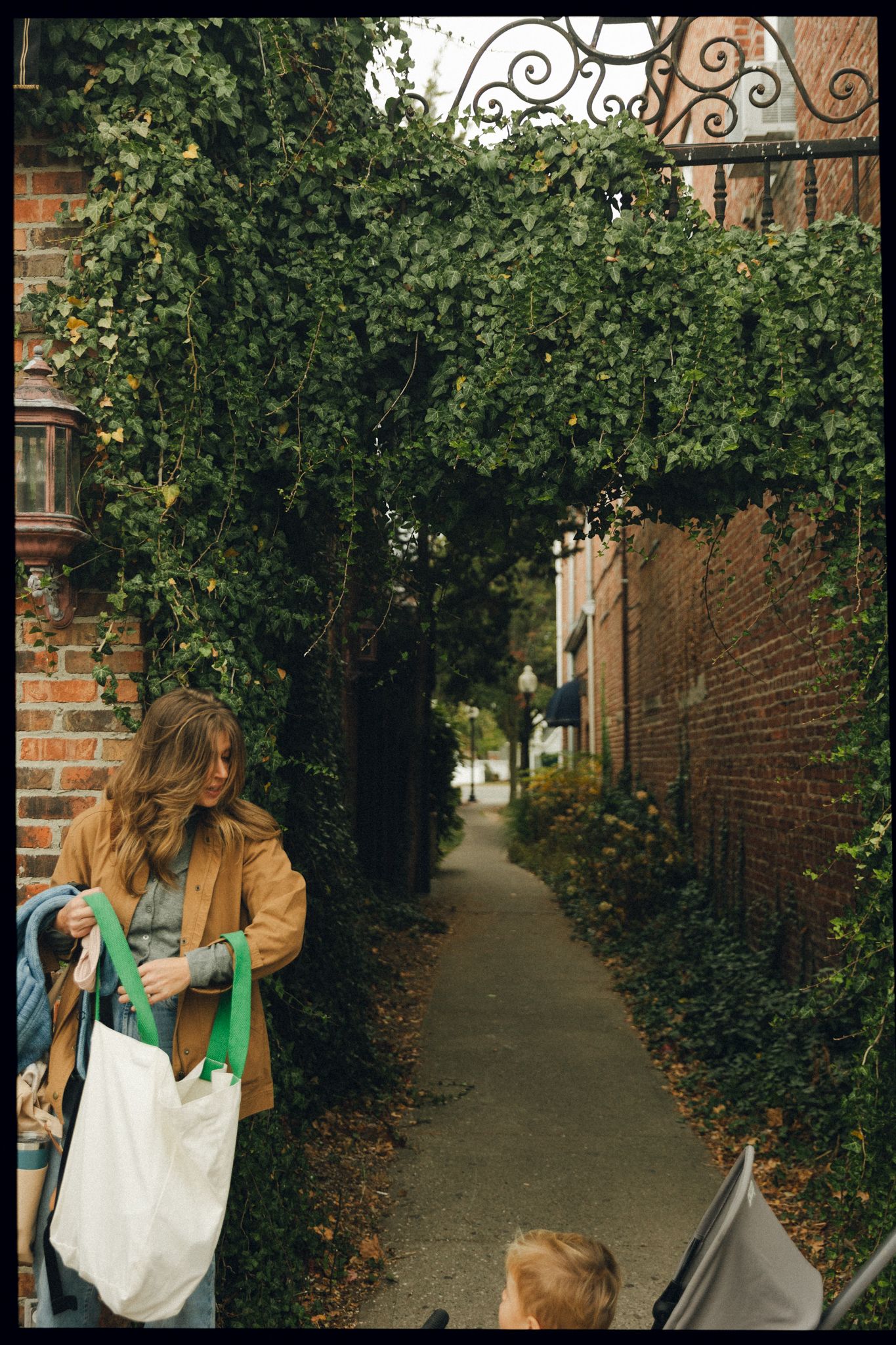 Woman in brown jacket carrying tote bag walks through ivy-covered brick archway on a charming narrow pathway.