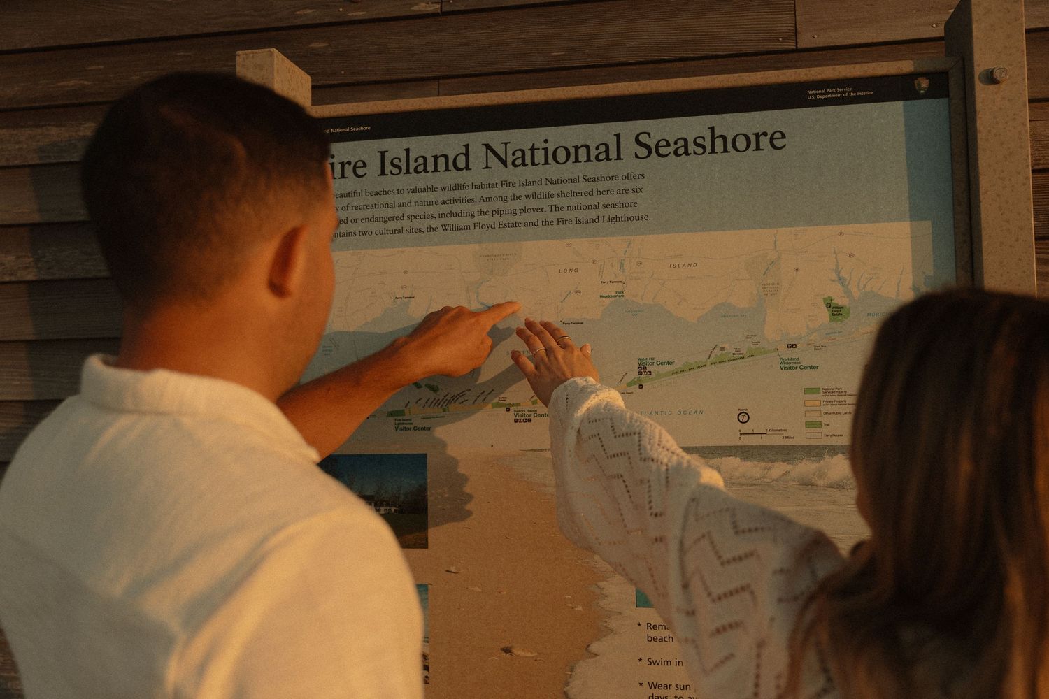 Couple pointing at Fire Island National Seashore map sign during golden hour sunset.