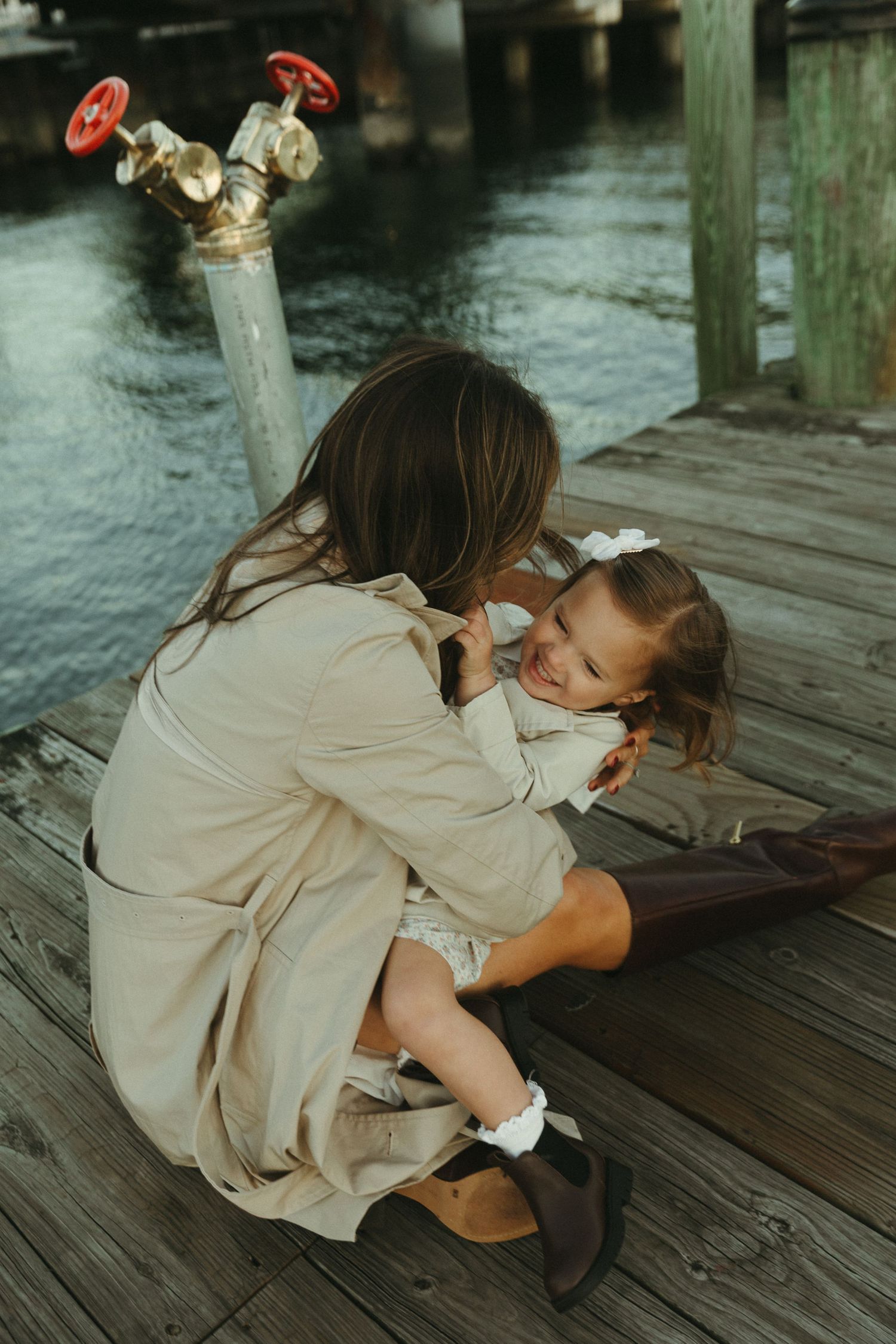 A person in a beige coat crouches down to examine something on a wooden dock.