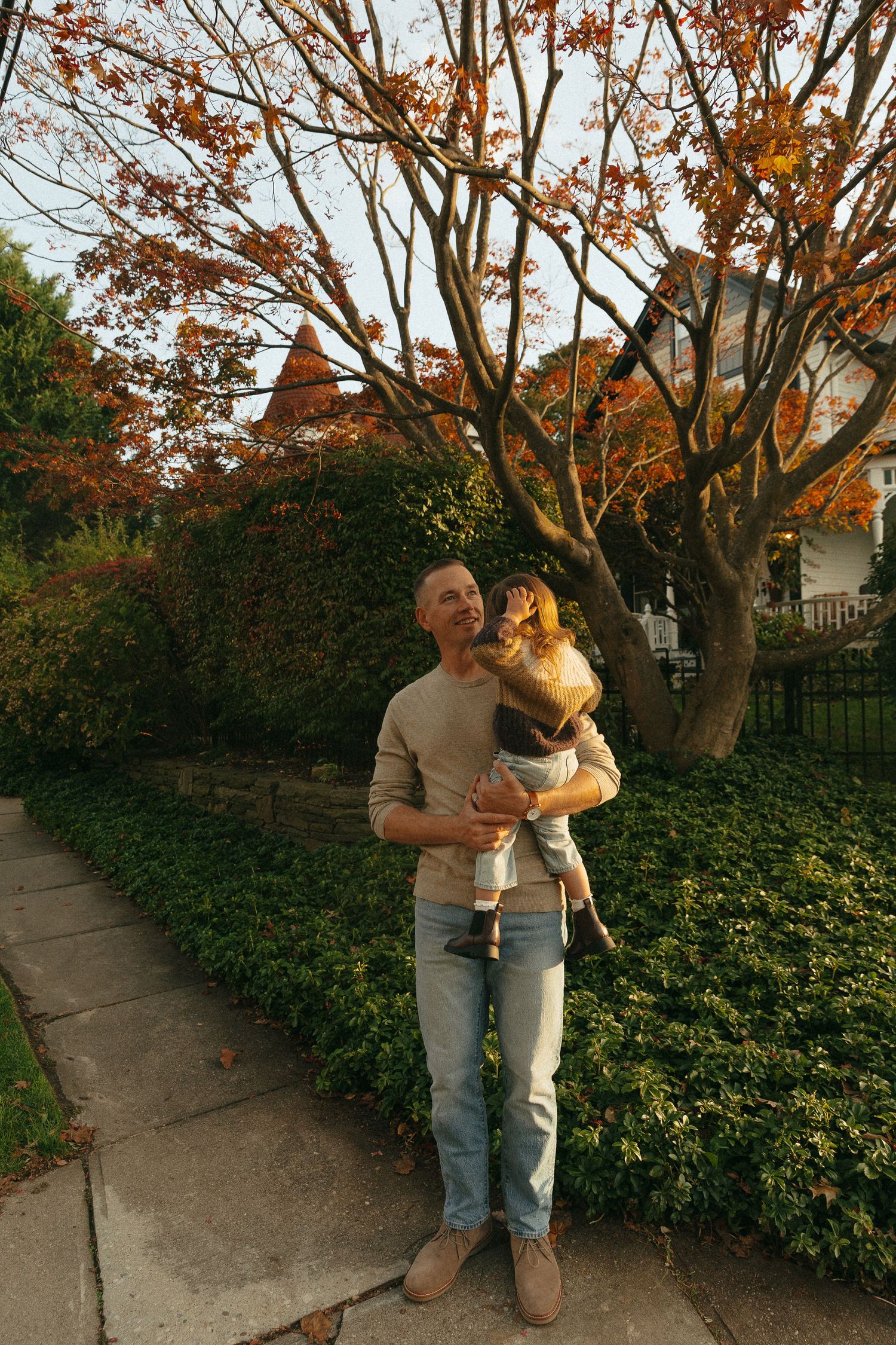 Man holding a young child outdoors on a sidewalk surrounded by autumn foliage and green hedges.
