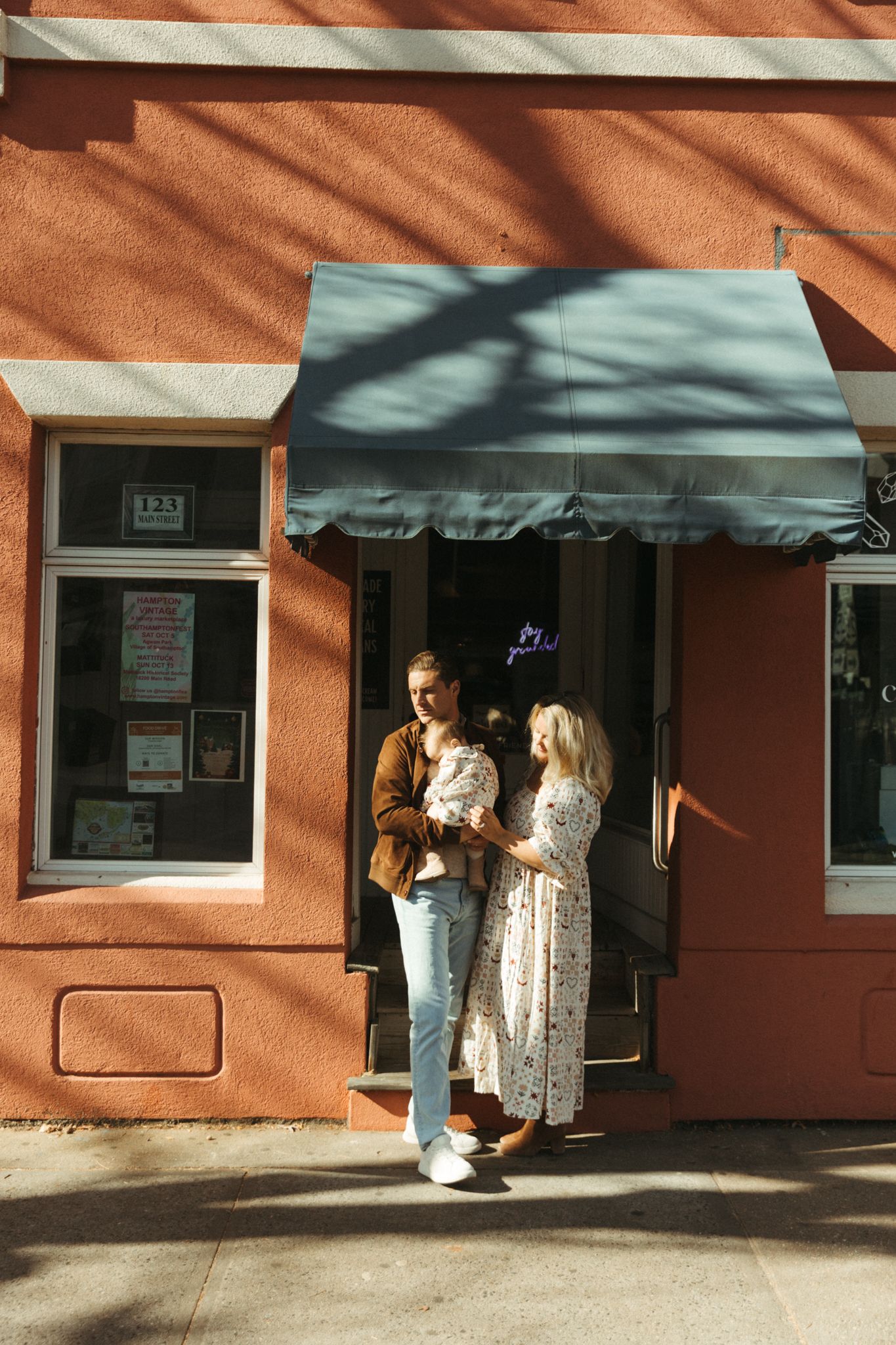 Couple with baby standing outside a red brick storefront with a teal awning on a sunny day.