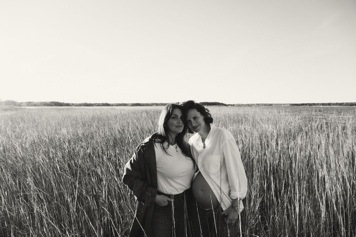 Black and white portrait of two women embracing in tall golden grass field at sunset.
